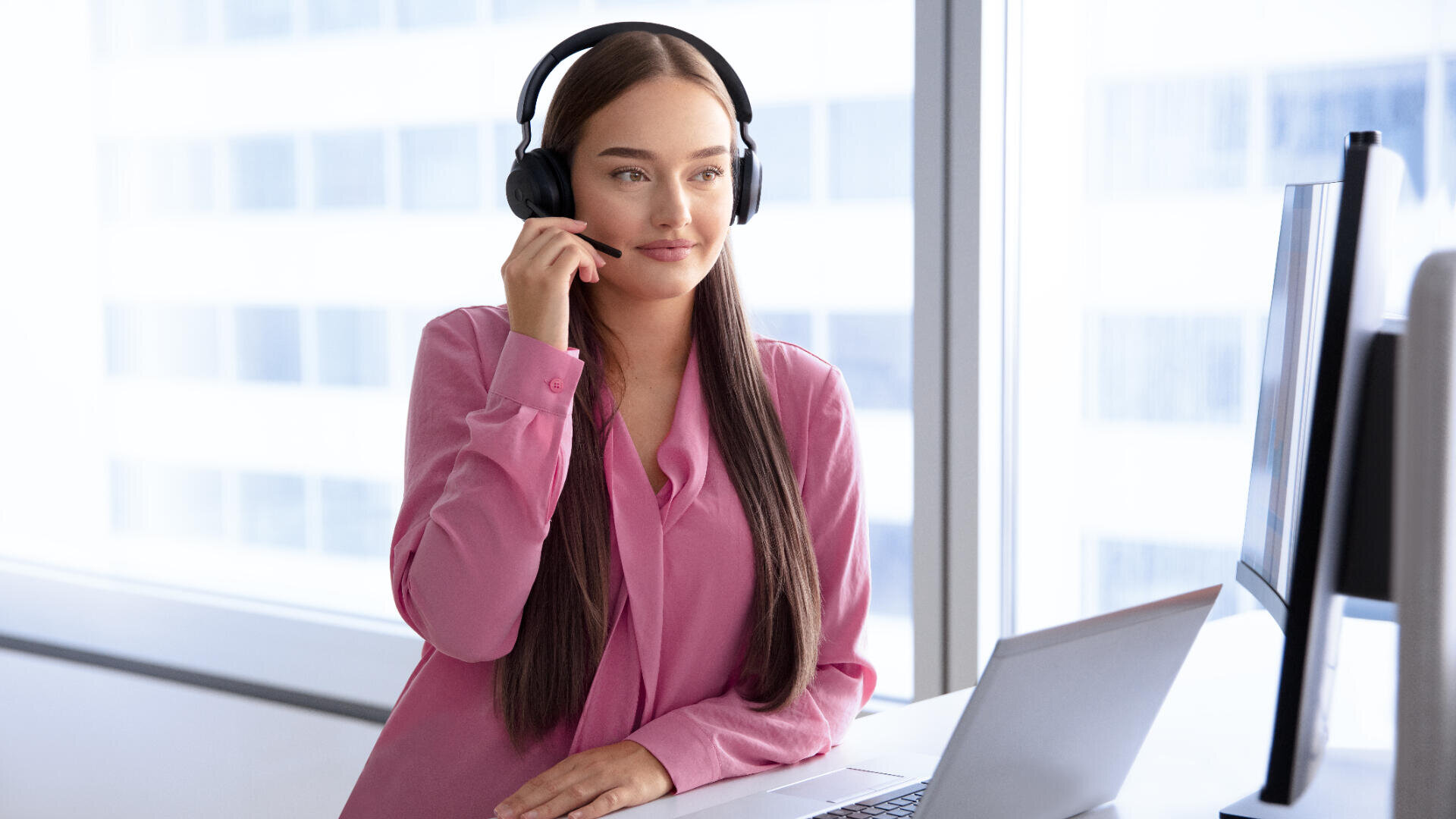 Eine Frau mit rosa Bluse und Headset sitzt an einem Schreibtisch mit Laptop, spricht und lächelt, während sie auf einen Computermonitor in einem hellen Büro mit großen Fenstern schaut.