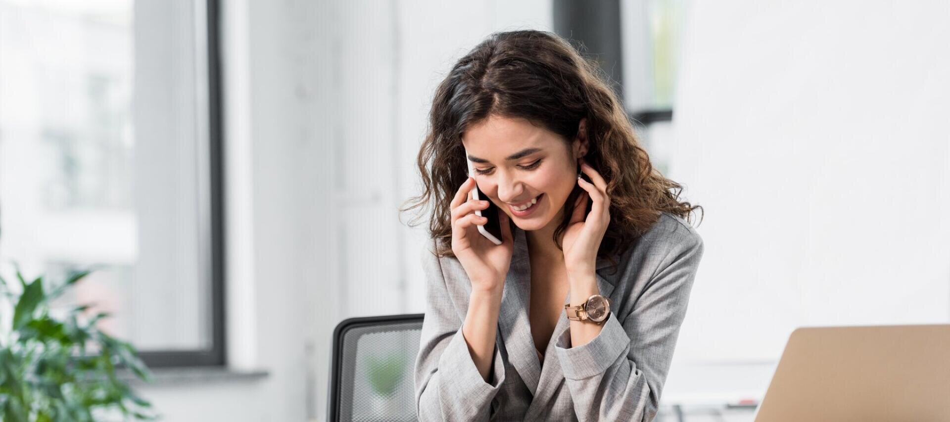 Eine lächelnde Frau mit lockigem Haar, die einen grauen Blazer trägt, sitzt an einem Schreibtisch und telefoniert mit ihrem Smartphone in einem hellen Büro.