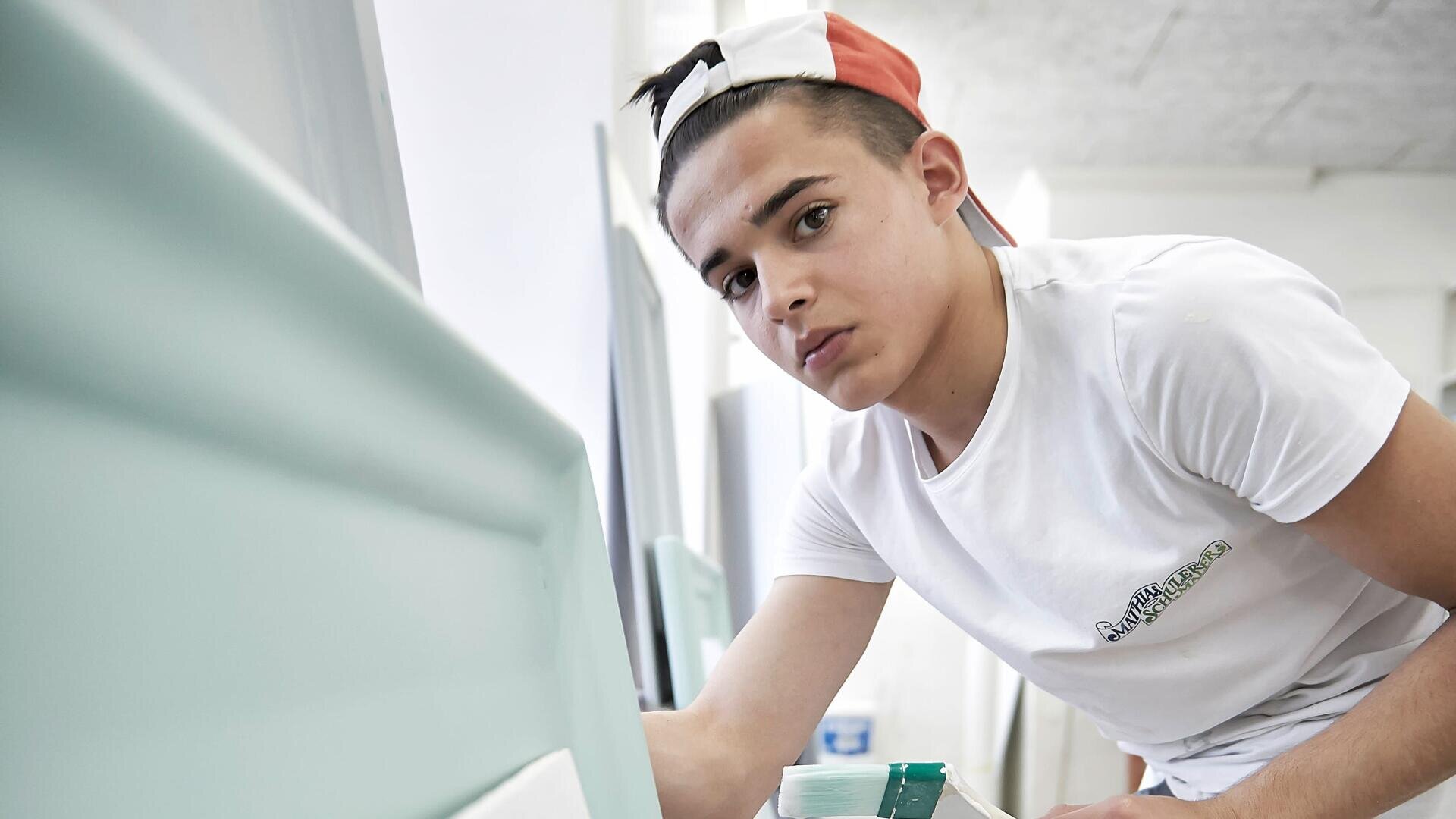 Eine junge Person mit weißem T-Shirt und umgekehrter Mütze streicht in einem gut beleuchteten Raum ein Möbelstück hellblau an und schaut dabei direkt in die Kamera.