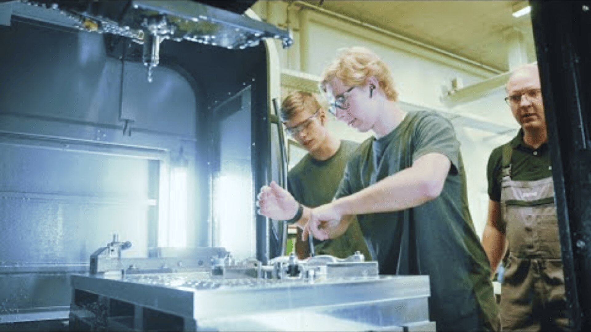 Drei Personen, die alle Schutzbrillen tragen, arbeiten gemeinsam in einer Industriehalle. Eine Person bedient die Maschinen, während die anderen genau beobachten. Die helle Beleuchtung hebt den Arbeitsbereich und die metallischen Oberflächen hervor.