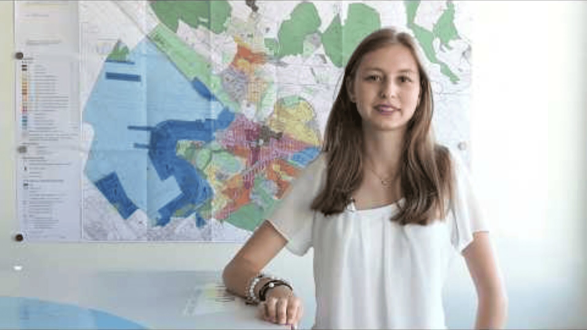 Eine junge Frau mit langen braunen Haaren und weißem Hemd steht lächelnd an einem Tisch, hinter ihr hängt ein bunter Stadtplan an der Wand.