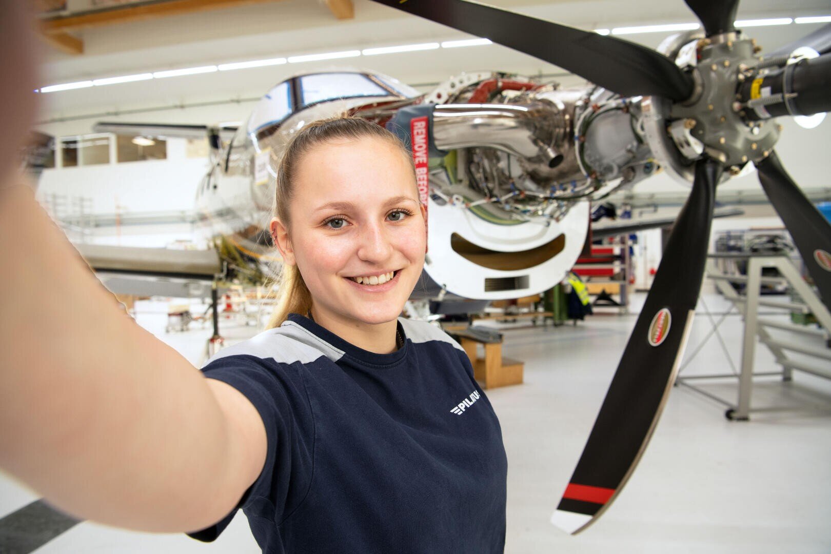 Eine Frau lächelt und macht ein Selfie vor einem Flugzeugtriebwerk in einem hell erleuchteten Hangar, im Hintergrund sind Maschinen und Werkzeuge zu sehen.