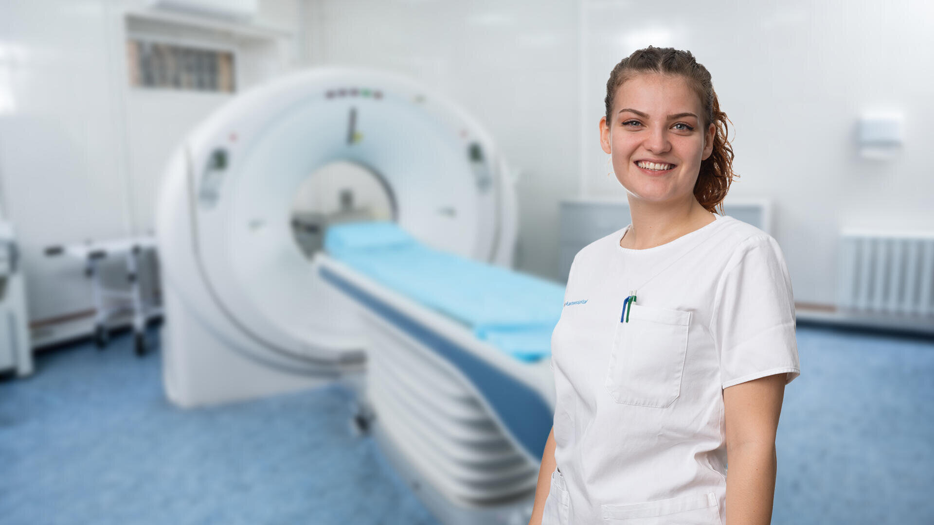 Eine lächelnde medizinische Fachkraft in weißem Kittel steht vor einem medizinischen Bildgebungsgerät, wahrscheinlich einem MRT- oder CT-Scanner, in einem sauberen, modernen Krankenhauszimmer.