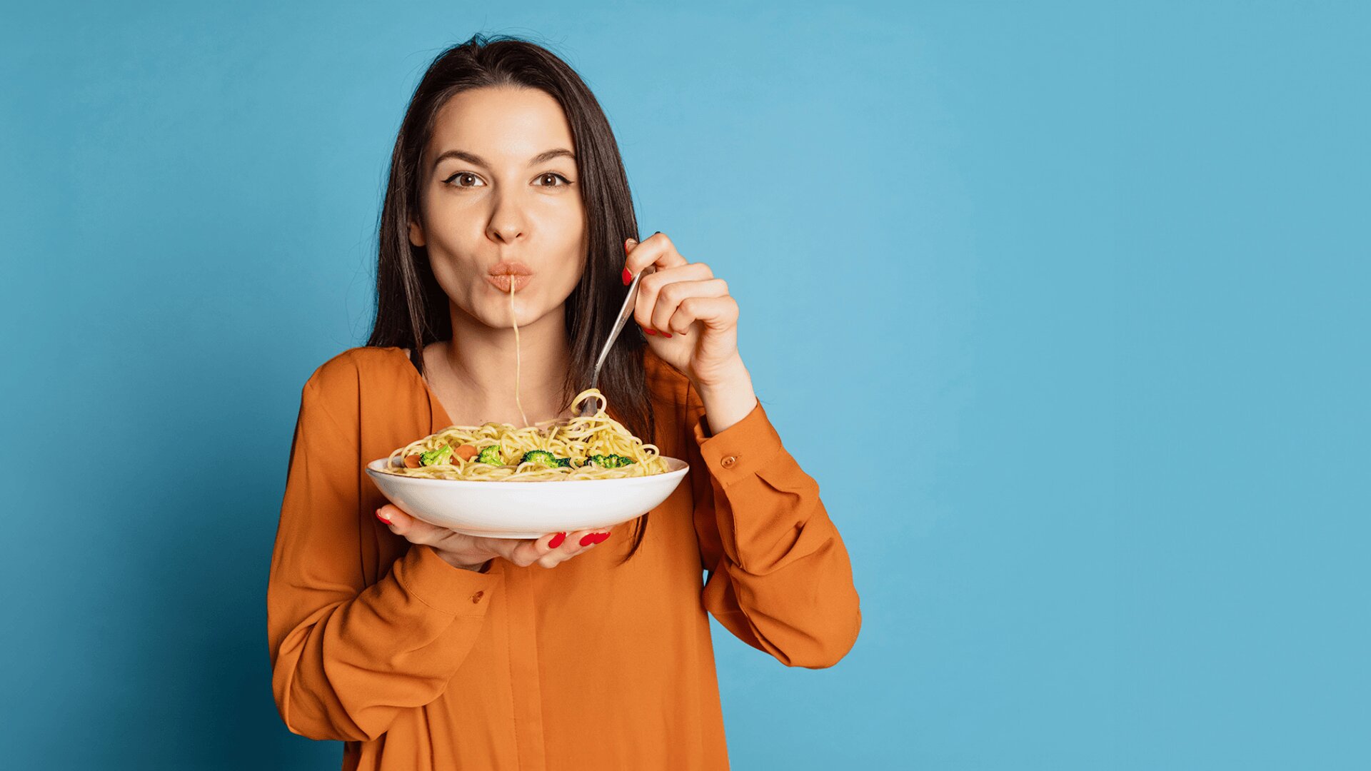 Eine Frau in orangefarbener Bluse hält eine Schüssel mit Spaghetti in der Hand und isst einen Nudelstrang vor einem einfarbig blauen Hintergrund.