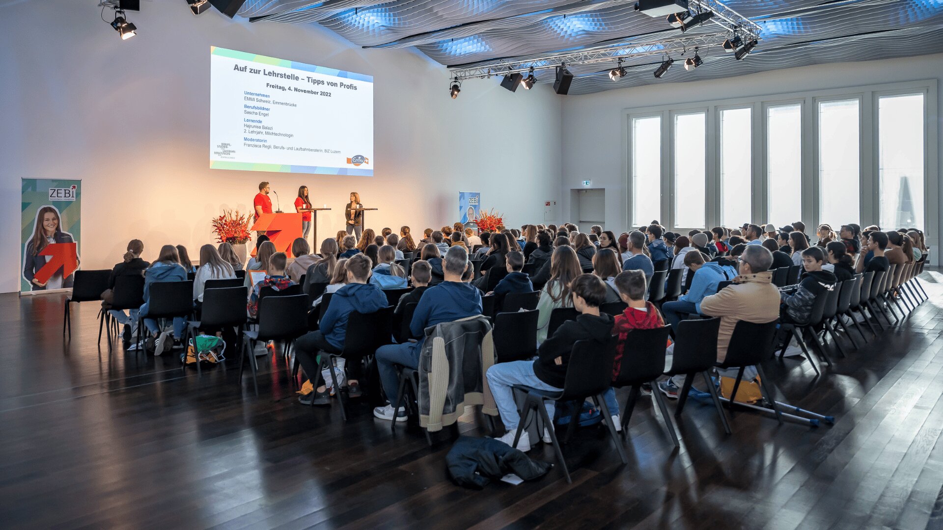 Eine große Gruppe von Personen sitzt in Reihen vor einer Bühne, auf der drei Redner in einem hellen, modernen Konferenzraum mit großen Fenstern Informationen auf einem Bildschirm präsentieren.