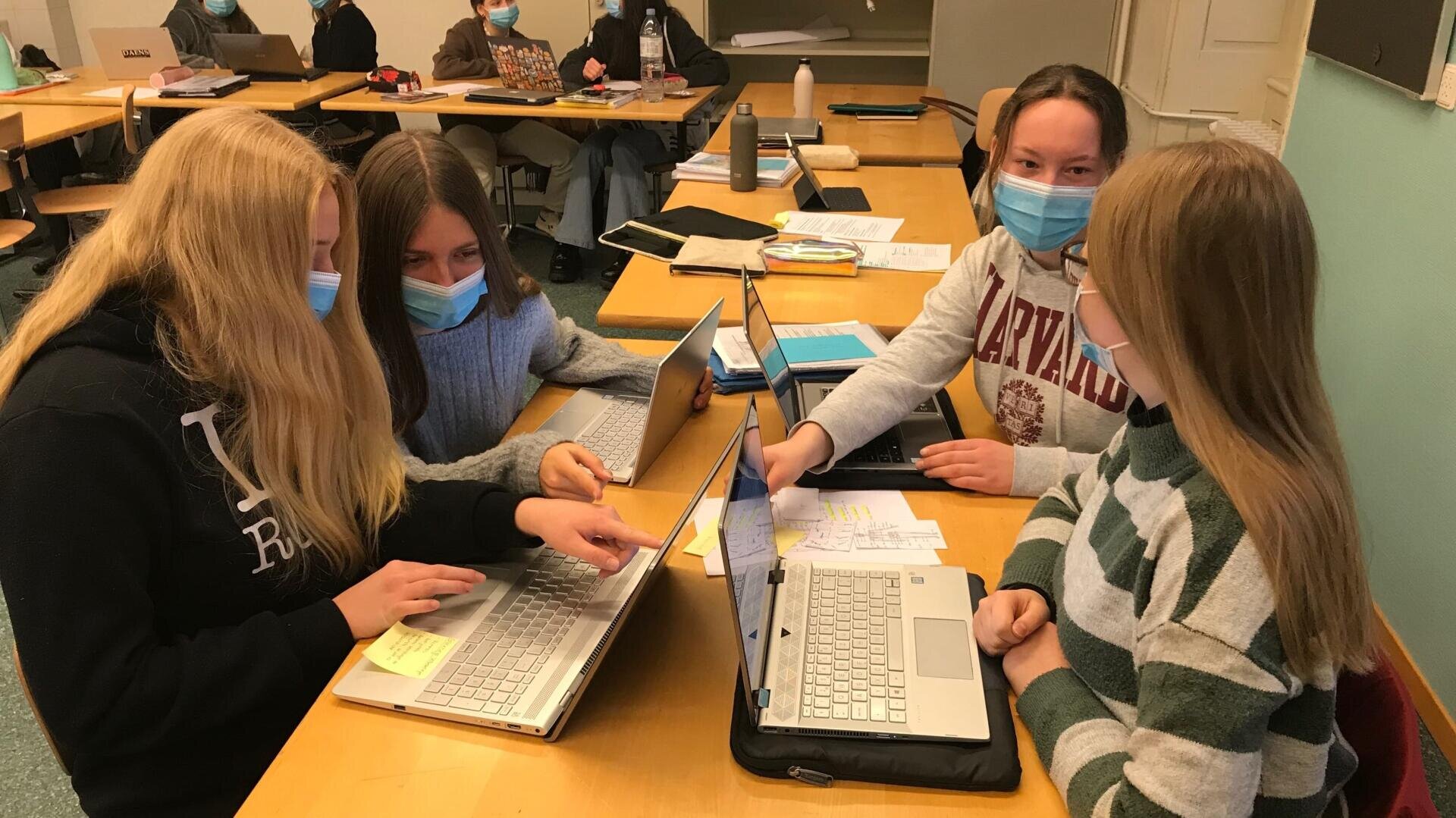 Vier maskierte Schüler sitzen an einem Tisch in einem Klassenzimmer und arbeiten gemeinsam an Laptops und Papieren. Im Hintergrund sind weitere Schüler zu sehen, die ebenfalls an Tischen arbeiten.