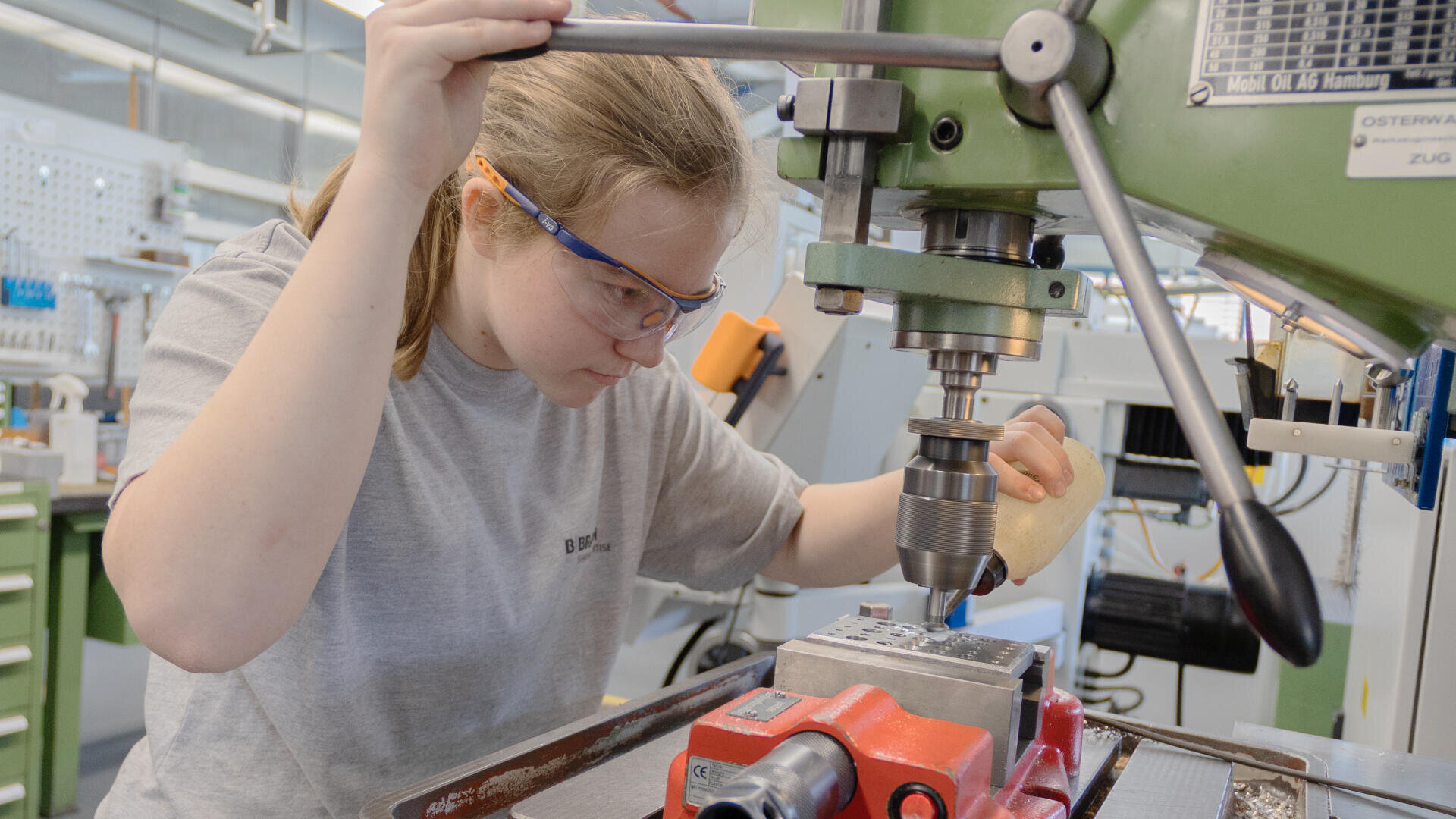 Eine Person, die eine Schutzbrille trägt, bedient eine grüne Industriebohrmaschine in einer Werkstatt. Sie konzentriert sich, während sie ein Werkstück auf der Maschine fixiert.