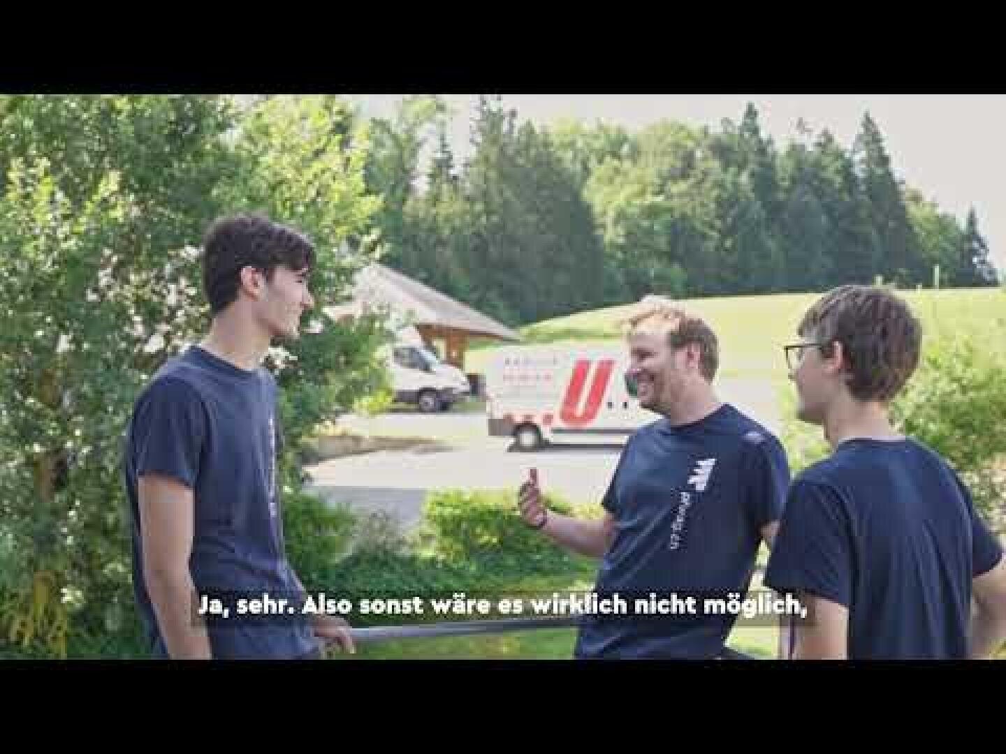 Drei junge Männer stehen an einem sonnigen Tag draußen, unterhalten sich und lachen, im Hintergrund sind Grünpflanzen und ein Lieferwagen zu sehen. Deutsche Untertitel sind am unteren Rand des Bildes zu sehen.