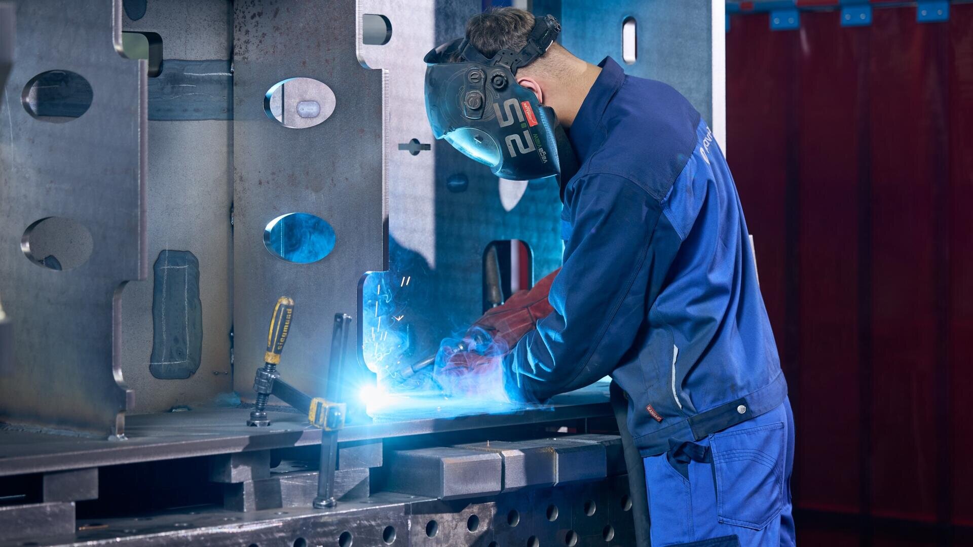 Ein Arbeiter, der einen Schweißhelm trägt, schweißt in einer Werkstatt an einer großen Metallkonstruktion.