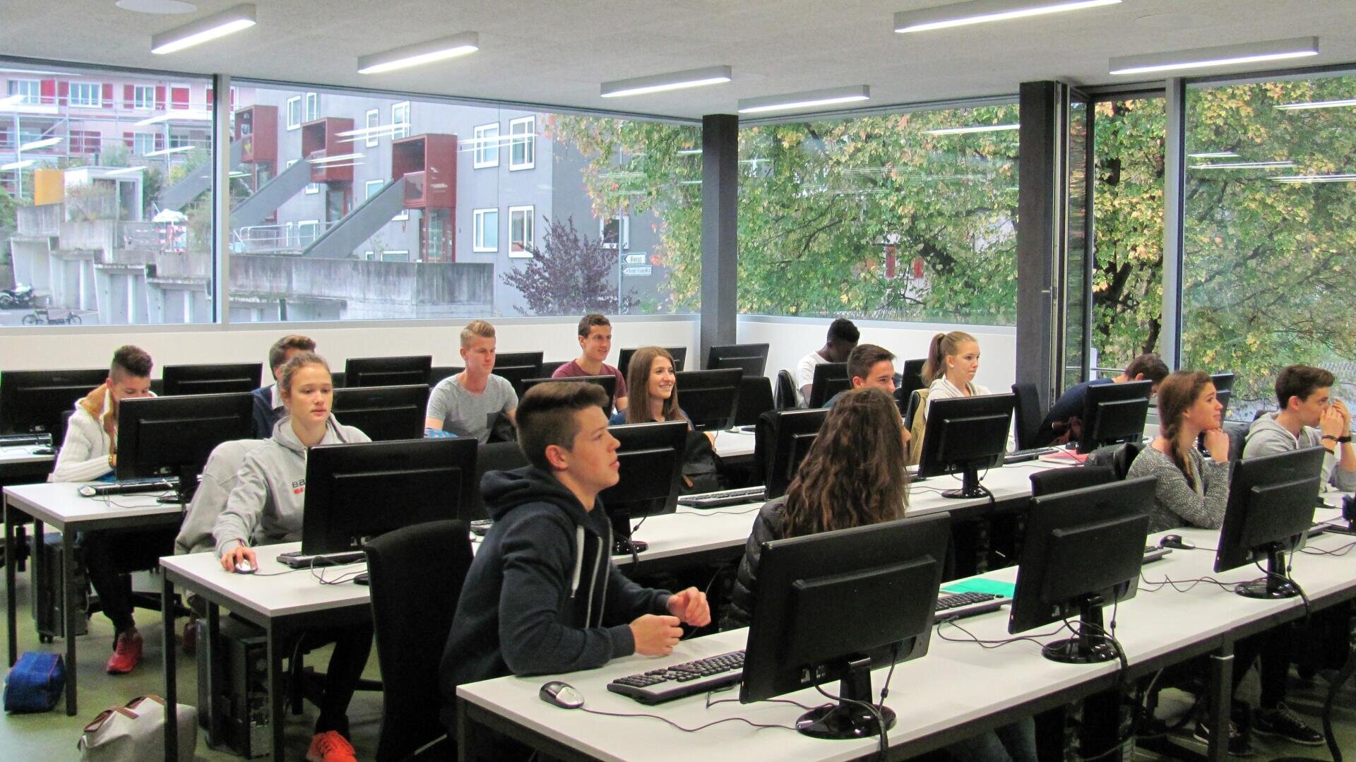 In einem Klassenzimmer sitzen die Schüler an Schreibtischreihen mit Desktop-Computern. Große Fenster geben den Blick auf Bäume und Gebäude frei. Die Schüler scheinen konzentriert zu sein, einige schauen auf die Bildschirme, andere hören zu.