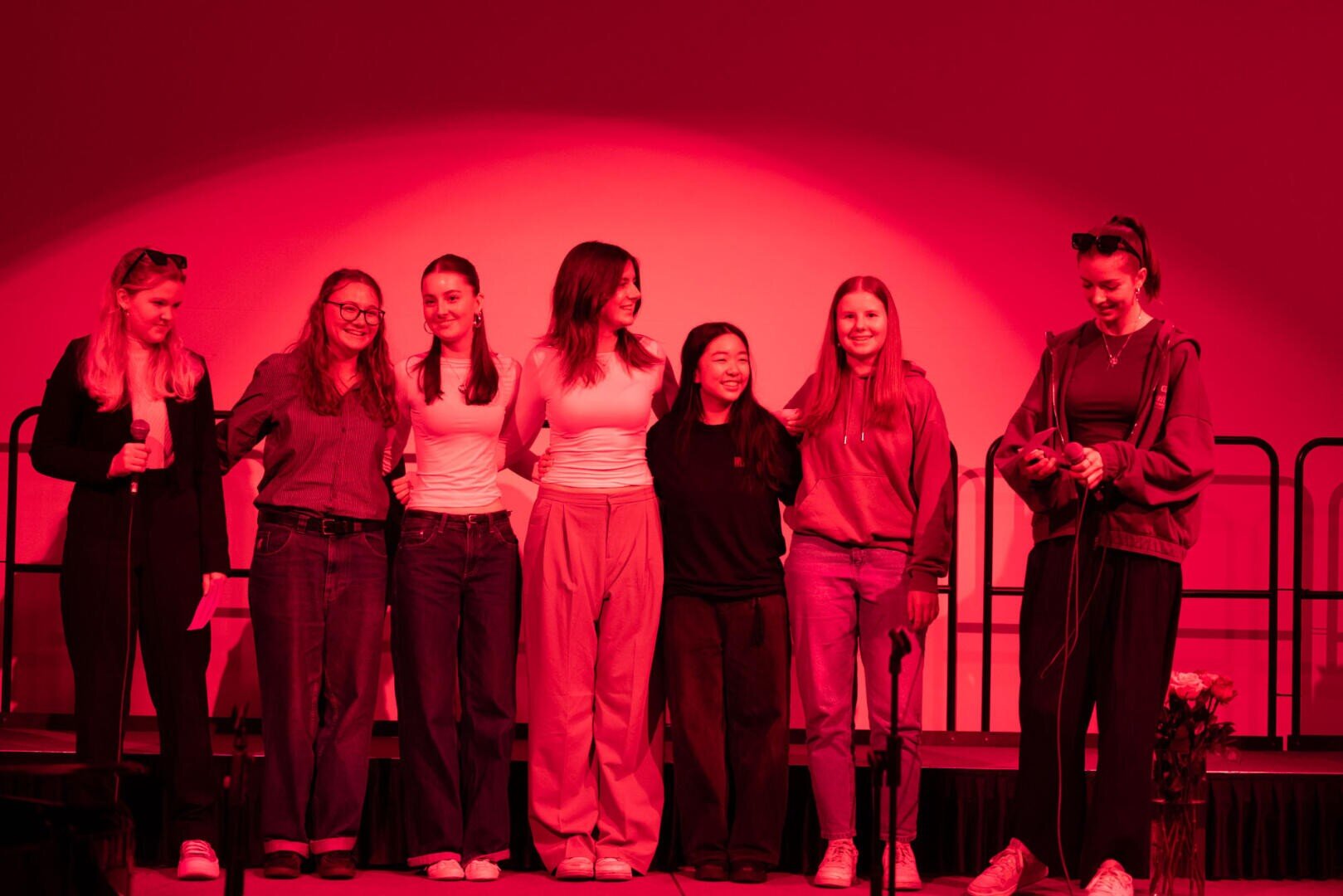 Sieben junge Frauen stehen zusammen auf der Bühne unter roter Beleuchtung, lächeln und posieren für ein Gruppenfoto. Einige haben die Arme umeinander gelegt. Ein Blumenstrauß liegt vor ihnen auf dem Boden.