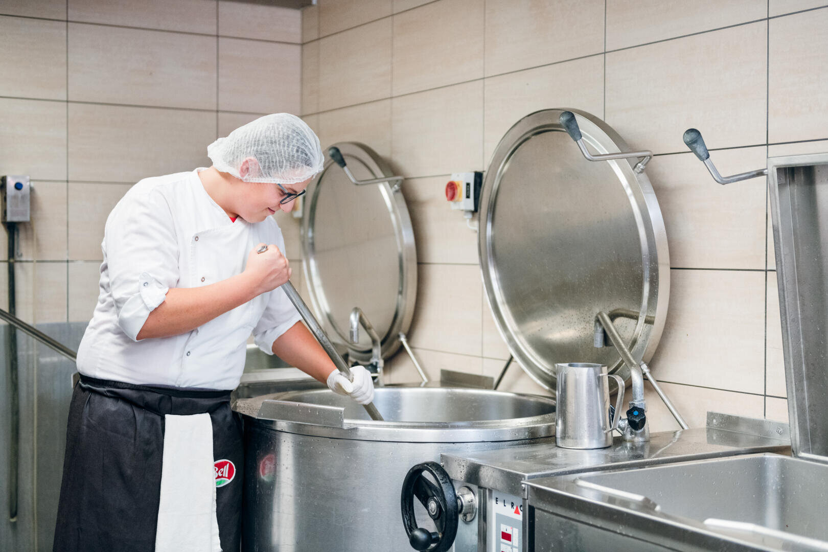 Ein Koch mit Haarnetz, Brille und weißer Uniform rührt in einer Großküche mit gefliesten Wänden und Geräten aus rostfreiem Stahl Speisen in einem großen Industrietopf.