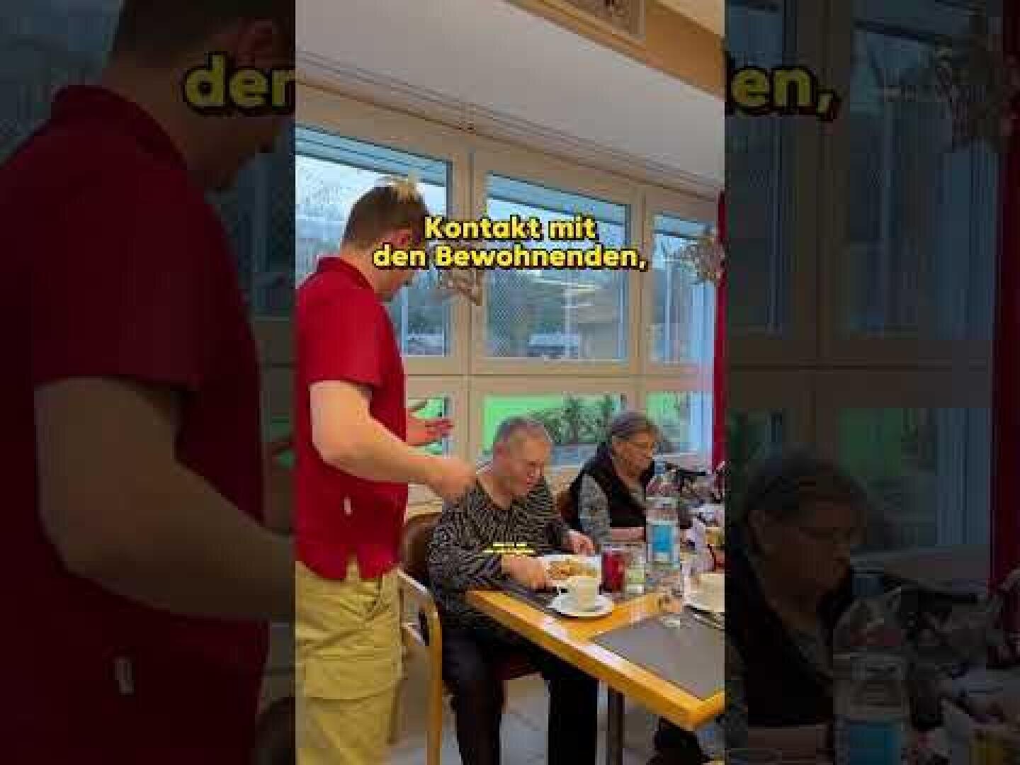 Eine Pflegekraft in einem roten Hemd steht neben sitzenden älteren Menschen an einem Esstisch in einem hellen Raum. Der deutsche Text auf dem Bild lautet "Kontakt mit den Bewohnenden", was so viel bedeutet wie "Kontakt mit den Bewohnern".