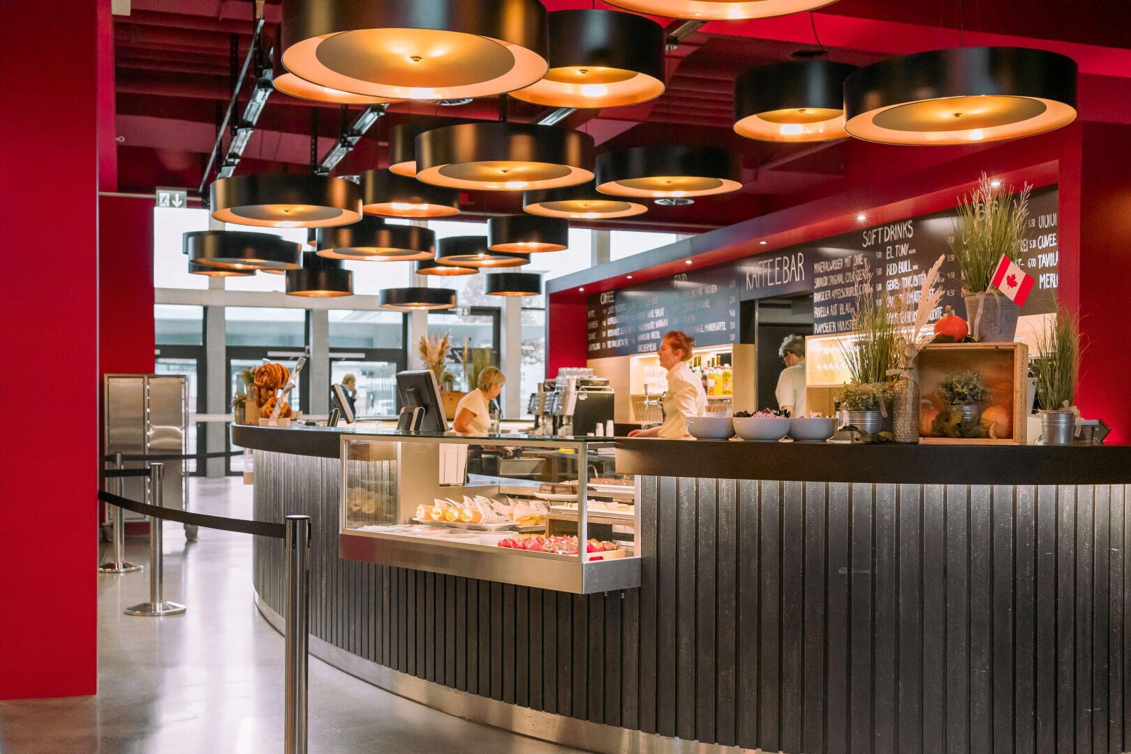 Modernes Café mit roten Wänden, kreisförmigen Hängelampen und einer geschwungenen Theke mit Gebäck. Das Personal arbeitet hinter dem Tresen, und im Hintergrund sind Menütafeln zu sehen. Einige Dekorationen und Pflanzen stehen auf dem Tresen.