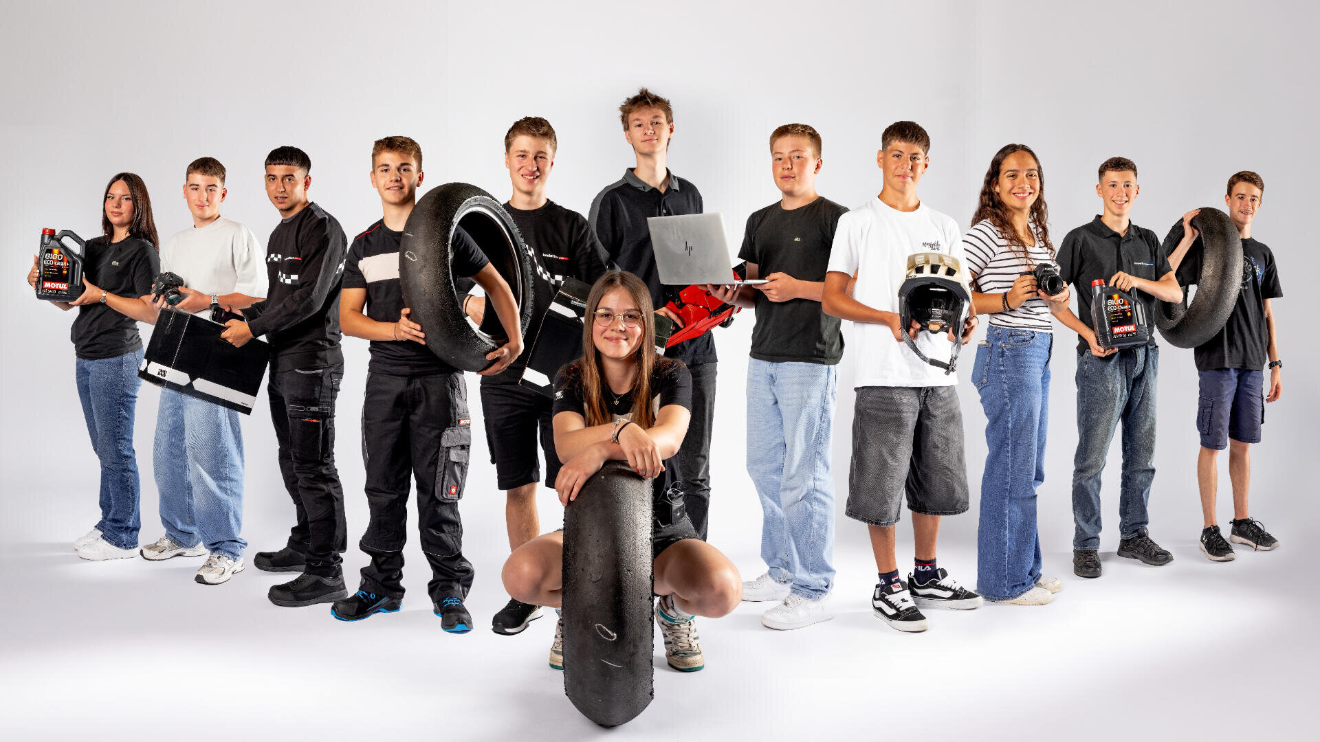 Eine Gruppe von zwölf Teenagern, von denen einige Rennreifen, Helme, Laptops oder Werkzeugkästen in der Hand halten, steht in einer Reihe vor einem weißen Hintergrund. Ein Mädchen kniet in der Mitte, lächelt und hält einen großen Reifen in der Hand.