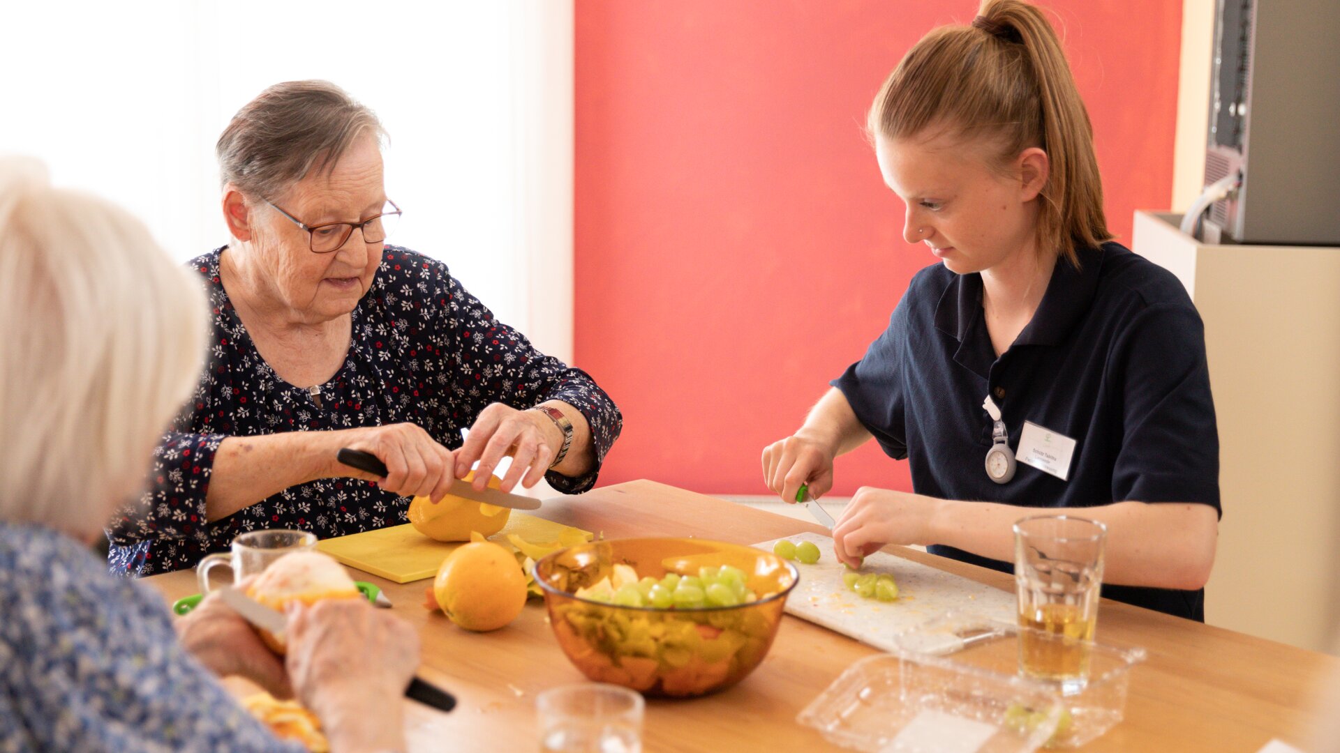 Zwei ältere Frauen und eine junge Betreuerin sitzen an einem Tisch und bereiten gemeinsam Obst zu, indem sie Zitronen und Trauben schneiden. Eine Schale mit gemischtem Obst und Gläser mit Wasser stehen auf dem Tisch in einem hell erleuchteten Raum.