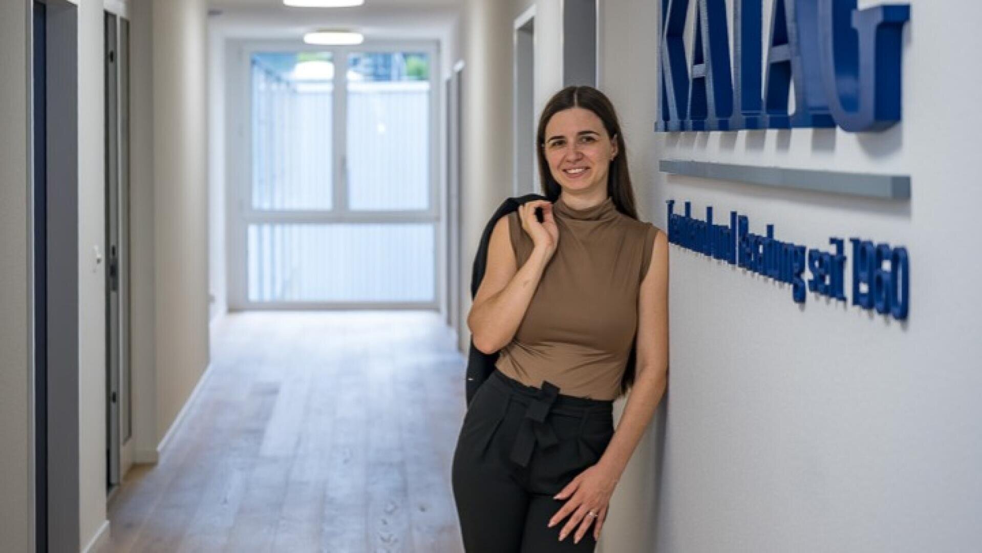 Eine Frau, die lächelnd an einer Wand lehnt, auf der das blaue Firmenlogo und der Schriftzug KATAG zu sehen sind, in einem hellen Büroflur.