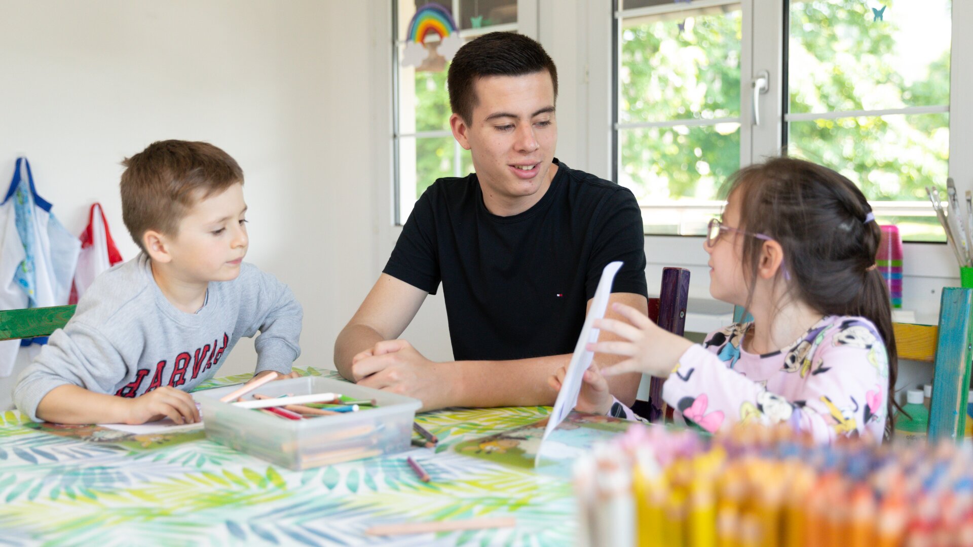 Ein junger Mann sitzt an einem Tisch mit einem Jungen und einem Mädchen, die zeichnen und sich unterhalten. Der Tisch ist mit Malutensilien bedeckt, und im Vordergrund liegen bunte Bleistifte. Sonnenlicht strömt durch die Fenster im Hintergrund.
