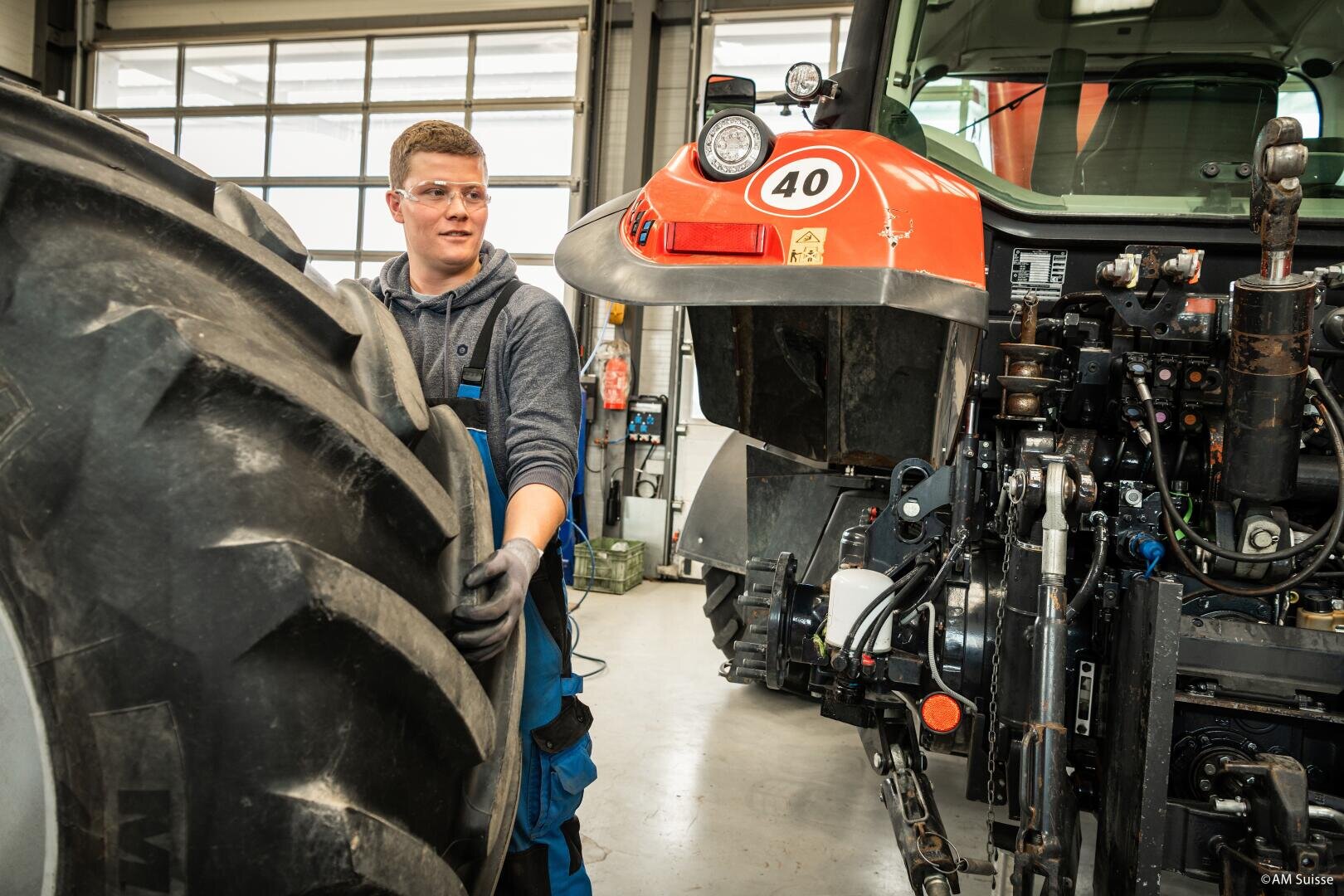 Ein Mann mit Schutzbrille und Handschuhen arbeitet in einer Werkstatt an einem großen Traktorreifen. Neben ihm ist das Heck eines roten Traktors mit der Nummer 40 zu sehen. Die Werkstatt ist mit großen Fenstern hell erleuchtet.