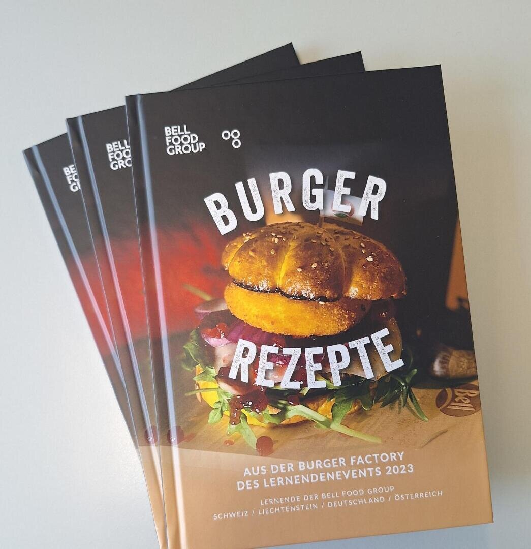 Ein Stapel Broschüren mit dem Titel Burger Rezepte und einem Foto eines Burgers auf dem Umschlag. Die Broschüre ist von der Bell Food Group für eine Veranstaltung im Jahr 2023, mit einem Text, der sich auf die Schweiz, Deutschland, Liechtenstein und Österreich bezieht.