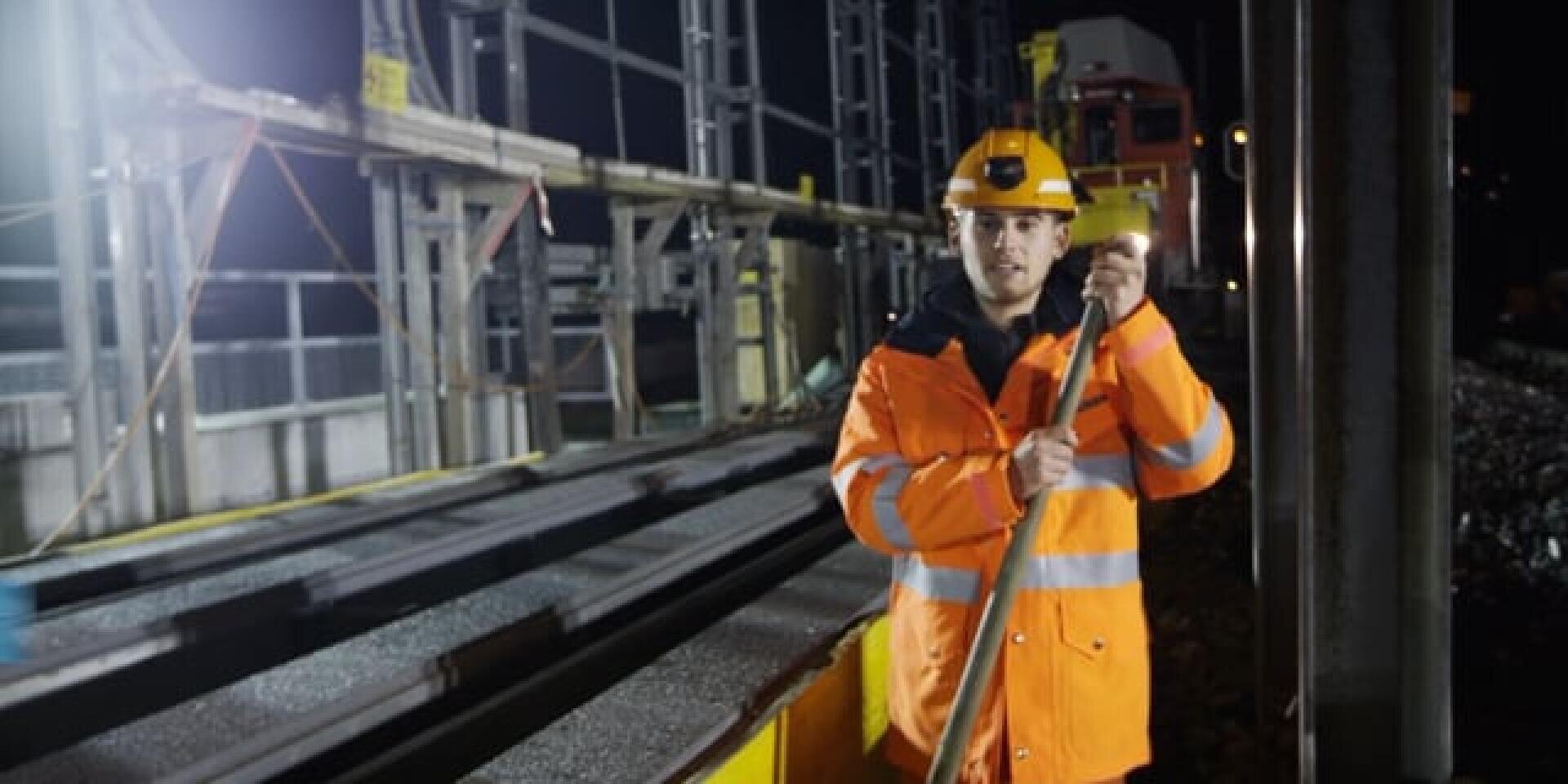 Ein Arbeiter in einer leuchtend orangefarbenen Sicherheitsweste und einem Schutzhelm steht nachts neben den Bahngleisen, hält ein Werkzeug in der Hand und blickt in die Kamera. Im Hintergrund sind Baumaschinen und Scheinwerfer zu sehen.