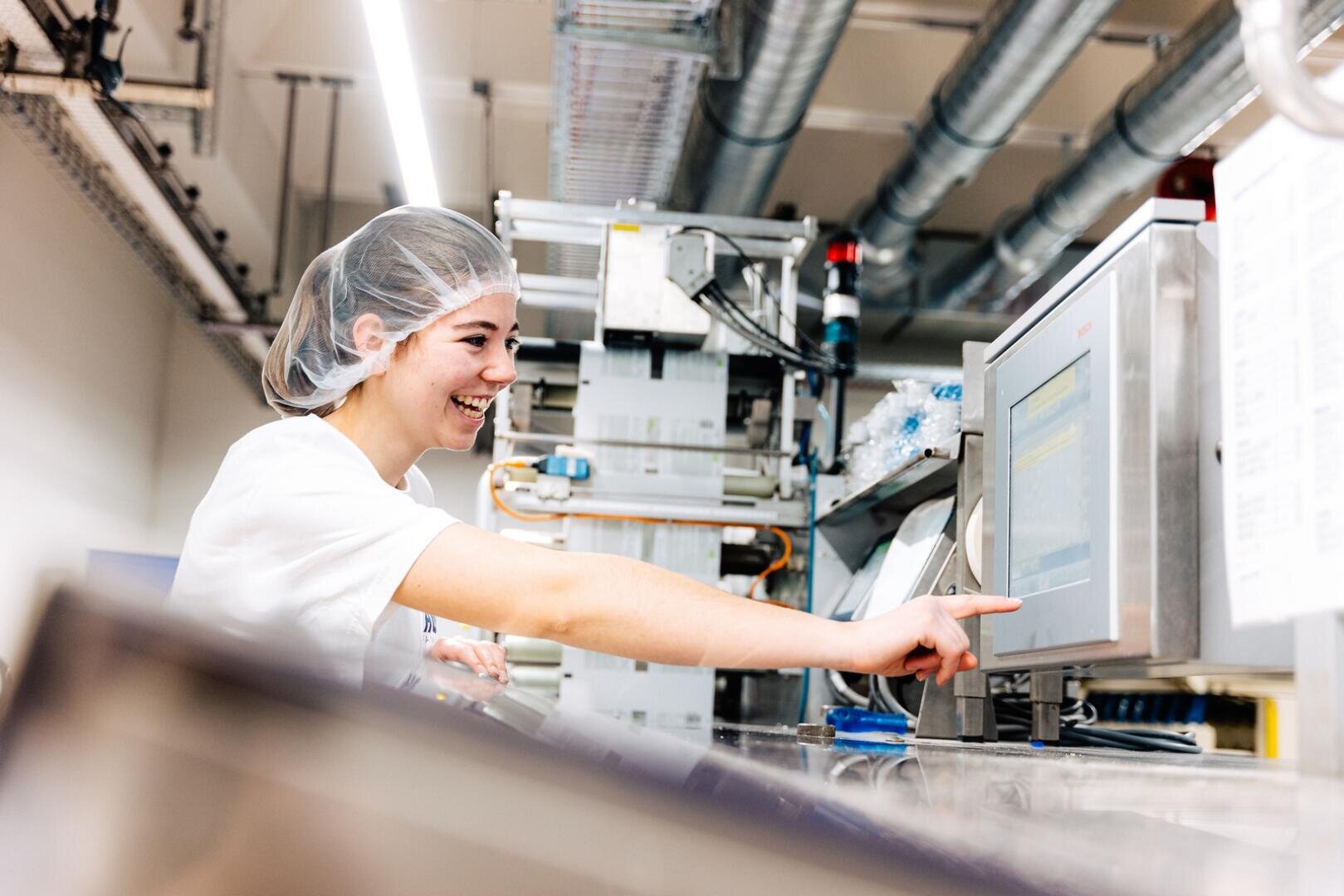 Eine Person, die ein Haarnetz und eine weiße Uniform trägt, lächelt, während sie in einer hell erleuchteten Fabrikumgebung einen Bildschirm einer Industrieanlage berührt.