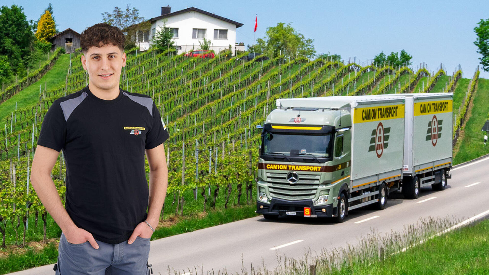 Ein junger Mann in einem schwarzen T-Shirt steht lächelnd vor einer Straße, auf der ein Camion Transport-LKW vorbeifährt, mit grünen Weinbergen und einem weißen Haus auf einem Hügel im Hintergrund.