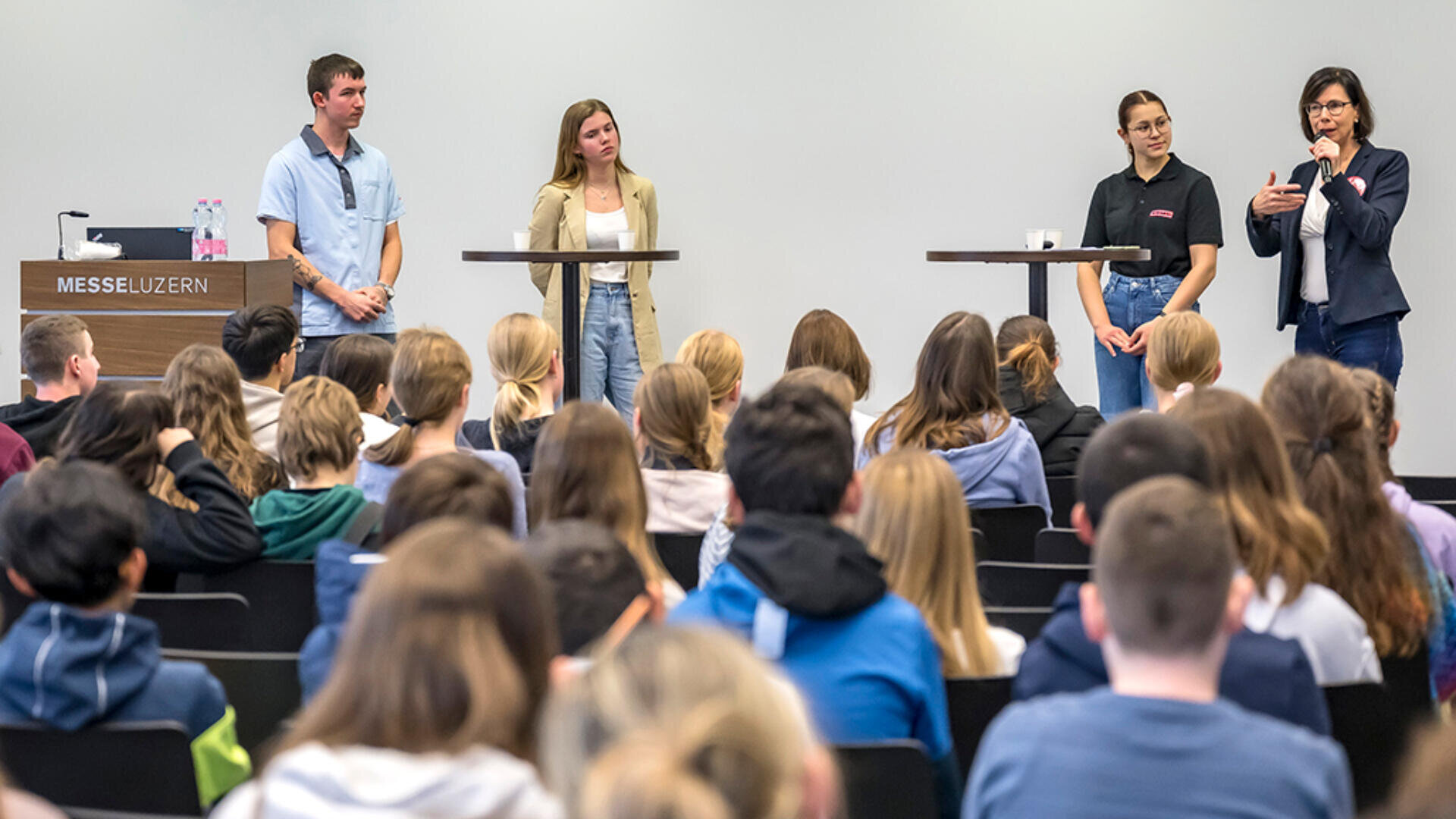 Vier Personen stehen vorne in einem Hörsaal und sprechen zu einem Publikum. Drei stehen hinter Podien, während eine mit der Hand gestikuliert und sich an die sitzende Menge wendet. Ein Schild mit der Aufschrift MESSE LUZERN steht auf einem Pult zur Linken.