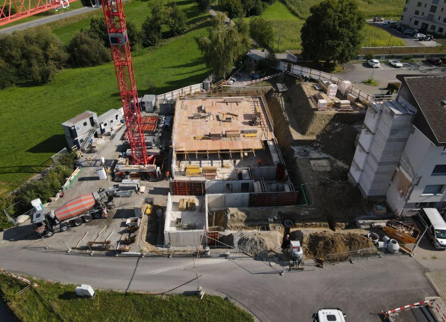 Luftaufnahme einer Baustelle mit einem großen roten Kran, Gebäudefundamenten, Baumaterialien, Fahrzeugen und Geräten, die ein teilweise gebautes Gebäude neben einer Straße und grünen Feldern umgeben.