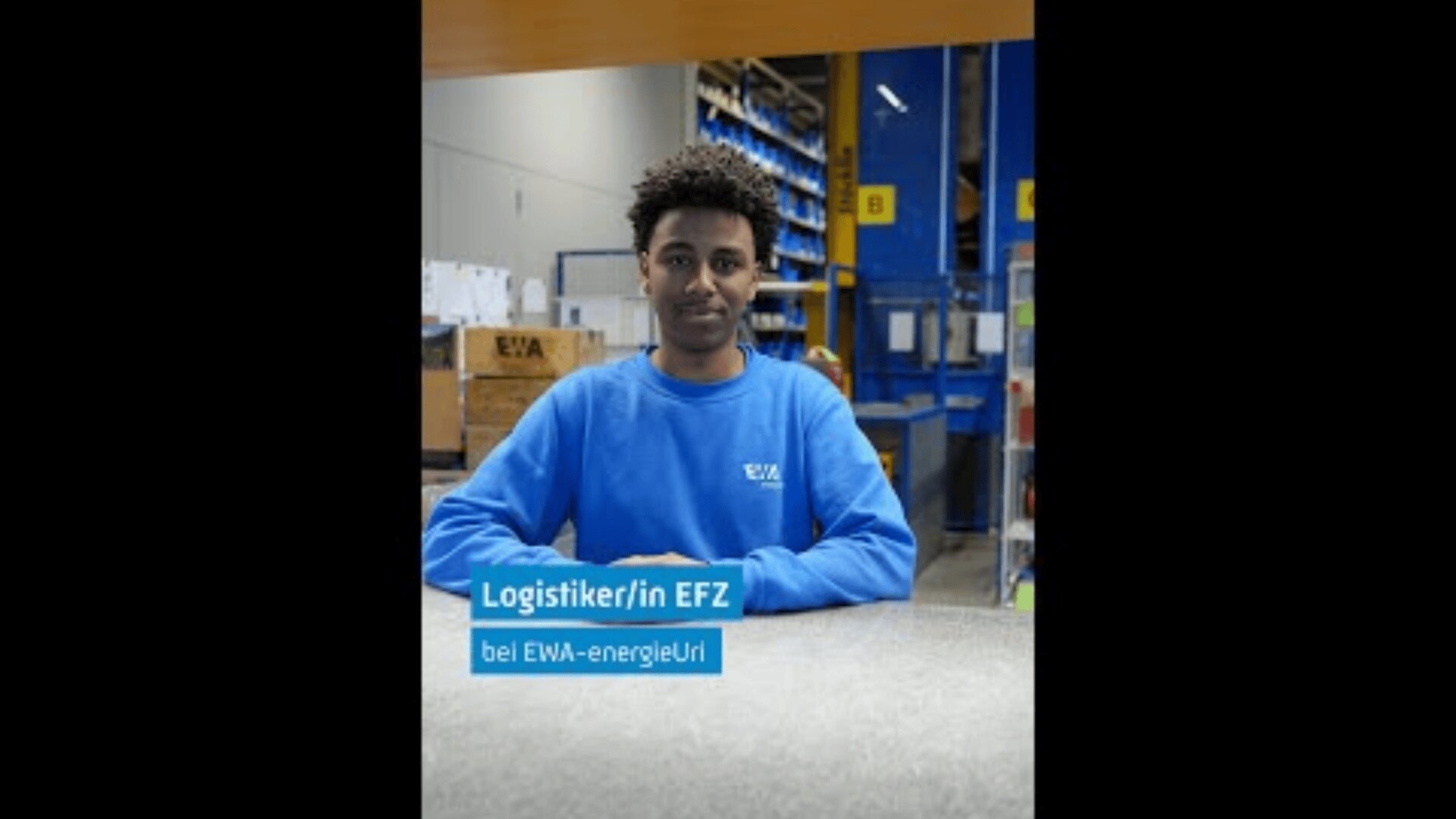 Eine Person mit kurzen lockigen Haaren und blauem EWA-energieUri-Hemd sitzt an einem Tisch in einer Lagerhalle mit Regalen und Kisten. Der Text lautet Logistiker/in EFZ bei EWA-energieUri.