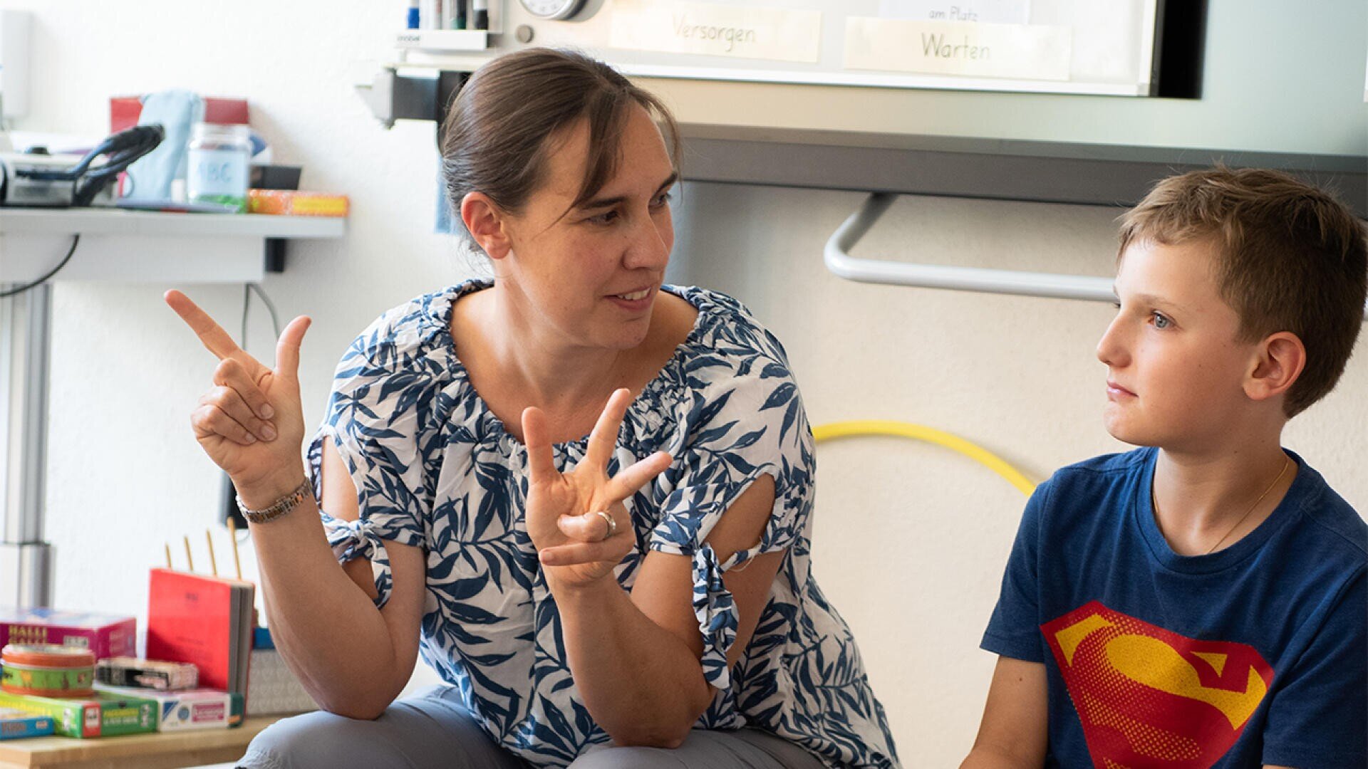 Eine Frau benutzt die Gebärdensprache, während sie neben einem kleinen Jungen in einem Klassenzimmer sitzt. Der Junge, der ein Superman-T-Shirt trägt, sieht sie aufmerksam an. Im Hintergrund sind Unterrichtsmaterialien und Kisten zu sehen.