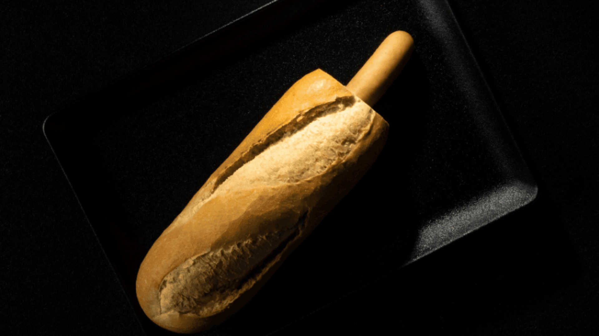 Ein Laib Brot mit einer in ein Ende gesteckten Brotstange auf einem schwarzen rechteckigen Tablett vor einem schwarzen Hintergrund.
