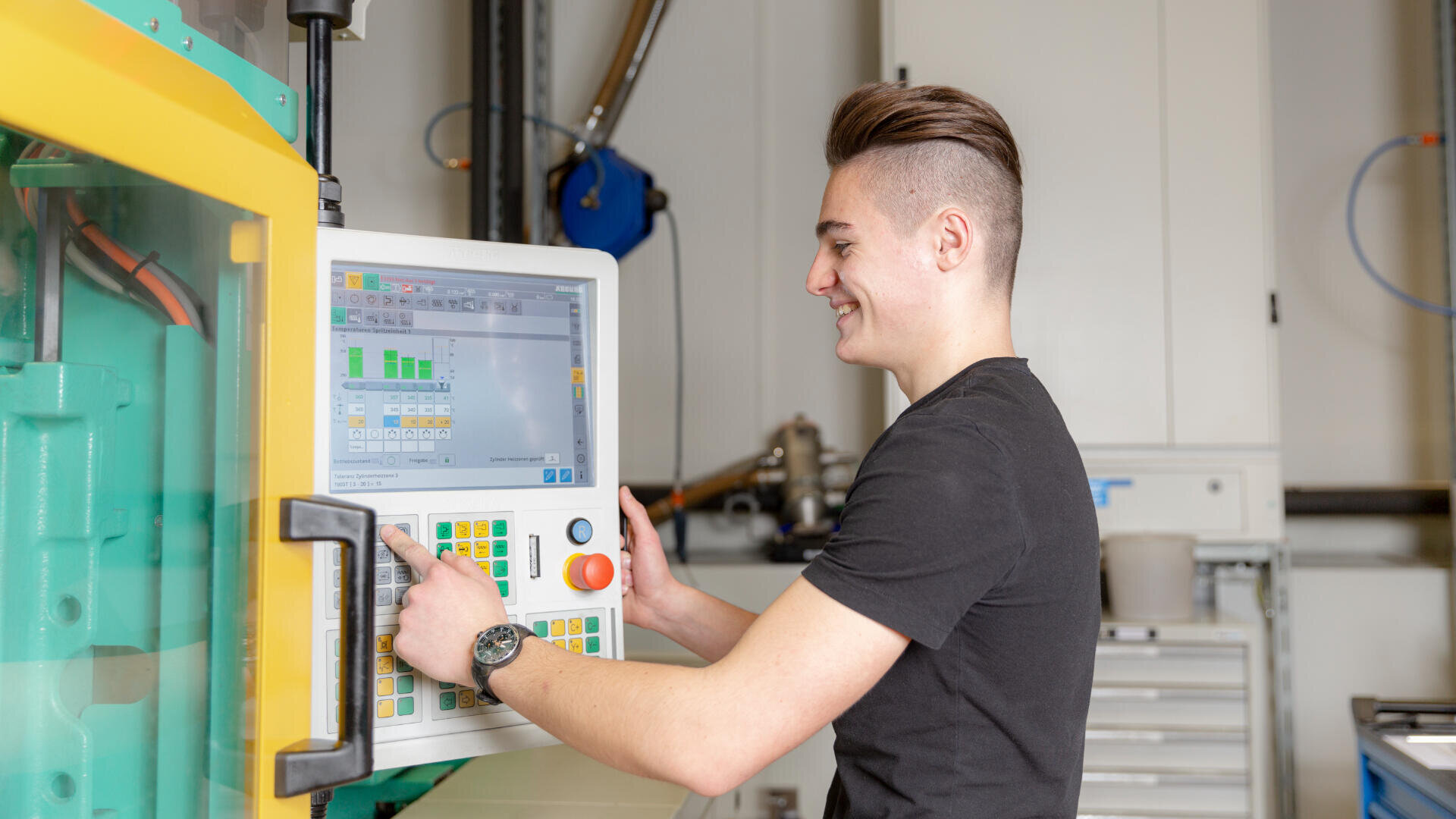 Ein junger Mann in einem schwarzen T-Shirt bedient in einer gut beleuchteten Werkstatt eine große Industriemaschine über ein digitales Bedienfeld mit Grafiken und Tasten.
