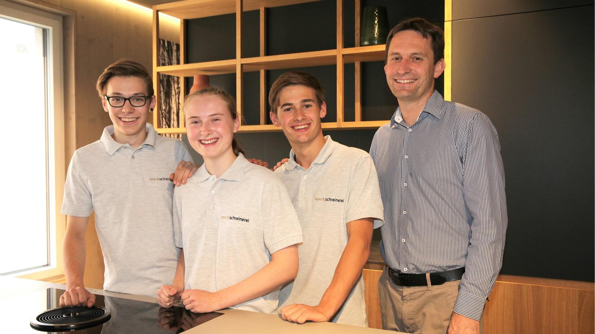 Drei lächelnde junge Leute in passenden hellgrauen Polohemden stehen neben einem Mann in einem gestreiften Button-up-Hemd. Sie alle posieren zusammen in einer modernen, gut beleuchteten Küche mit Holzregalen im Hintergrund.