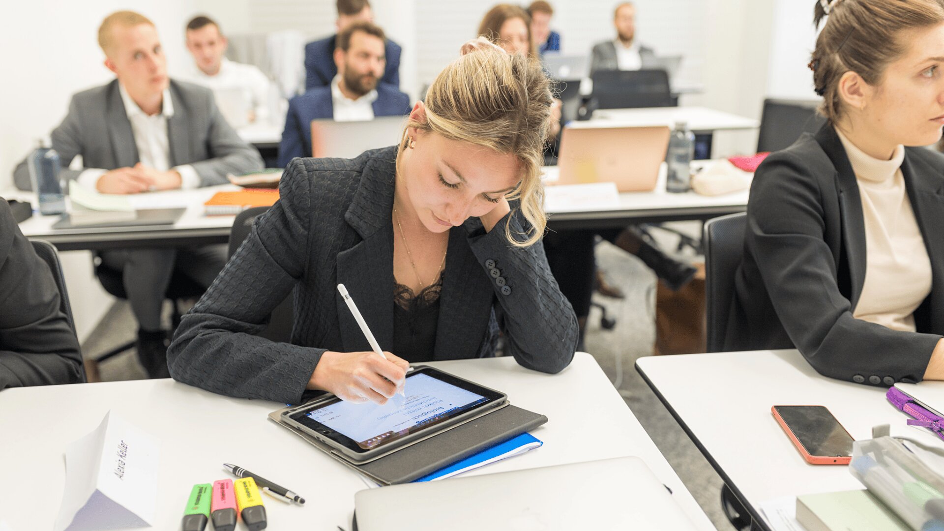Eine Frau in Business-Kleidung schreibt an einem Schreibtisch in einem Klassenzimmer auf einem Tablet, umgeben von anderen Erwachsenen, die zuhören und an Laptops und Tablets arbeiten. Auf den Tischen liegen Textmarker und Notizbücher.