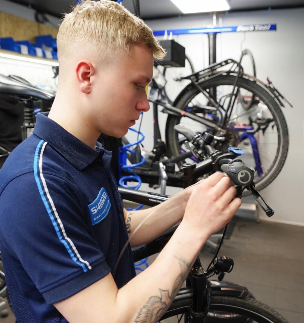 Ein junger Mann in einem blauen Shimano-Hemd repariert einen Fahrradlenker in einer Werkstatt, während im Hintergrund Werkzeuge und andere Fahrräder zu sehen sind.