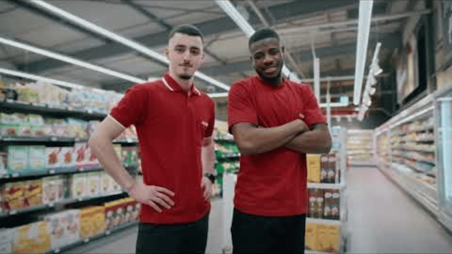 Zwei Angestellte eines Lebensmittelgeschäfts in roten Hemden stehen in einem Gang, lächeln und posieren für die Kamera. Im Hintergrund sind im hellen Licht des Ladens die mit Produkten gefüllten Regale zu sehen.