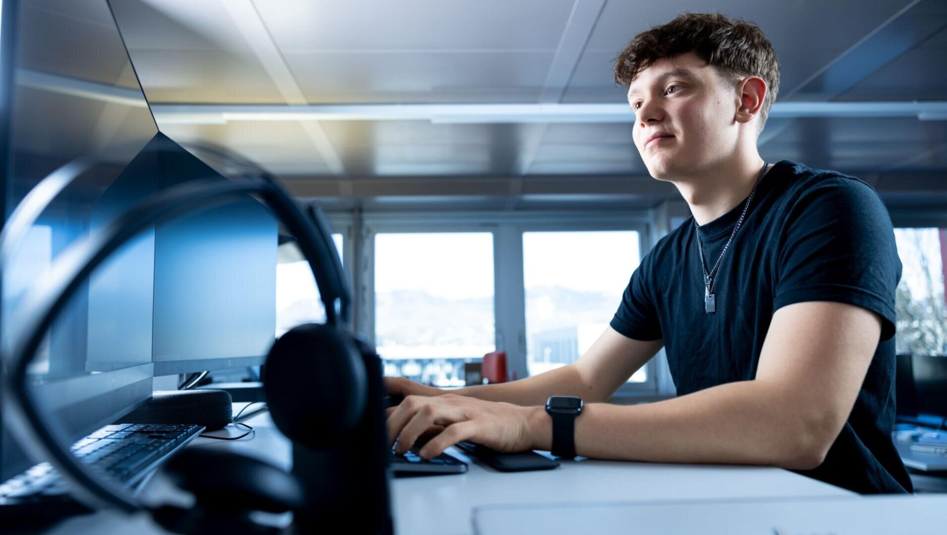 Ein junger Mann mit schwarzem Hemd und Smartwatch sitzt an einem Schreibtisch, tippt auf einer Tastatur und schaut auf einen Computermonitor in einem hellen, modernen Büro. Die Kopfhörer liegen auf dem Schreibtisch im Vordergrund.