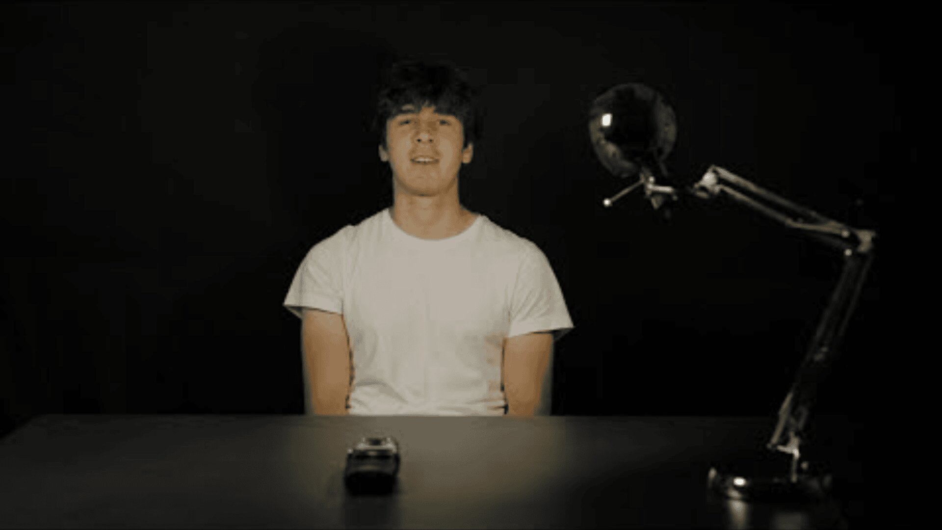 Eine junge Person in einem weißen T-Shirt sitzt an einem dunklen Tisch mit einem Tonbandgerät und einer Schräglichtlampe vor einem schwarzen Hintergrund.