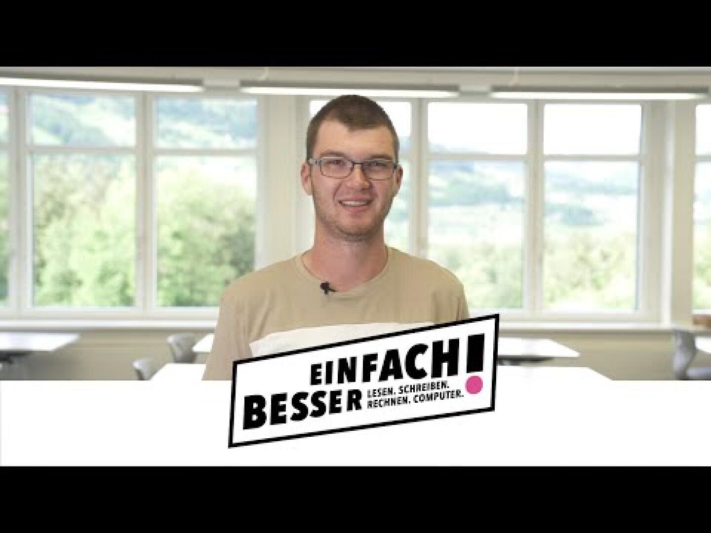 Ein junger Mann mit Brille steht lächelnd in einem hellen Klassenzimmer mit großen Fenstern. Davor steht ein Schild mit der Aufschrift EINFACH! BESSER LESEN. SCHREIBEN. RECHNEN. COMPUTER.