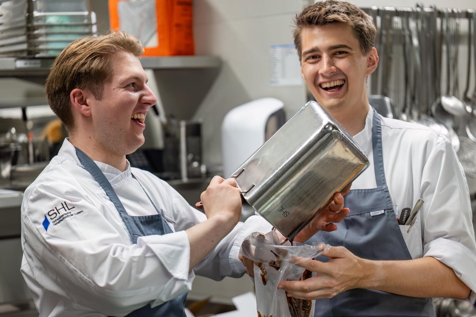 Zwei junge männliche Köche in weißen Uniformen und Schürzen lachen zusammen in einer Küche, während sie Schokoladenteig aus einem großen Metallbehälter in einen Spritzbeutel füllen. Im Hintergrund sind Küchengeräte und Utensilien zu sehen.