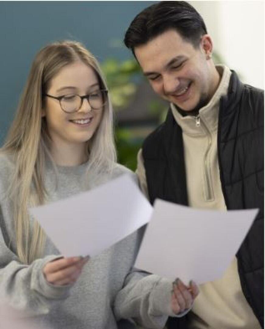 Zwei junge Erwachsene, einer mit langen blonden Haaren und Brille und einer mit kurzen dunklen Haaren, stehen nebeneinander, lächeln und halten Blätter in der Hand, als würden sie gemeinsam Dokumente prüfen.