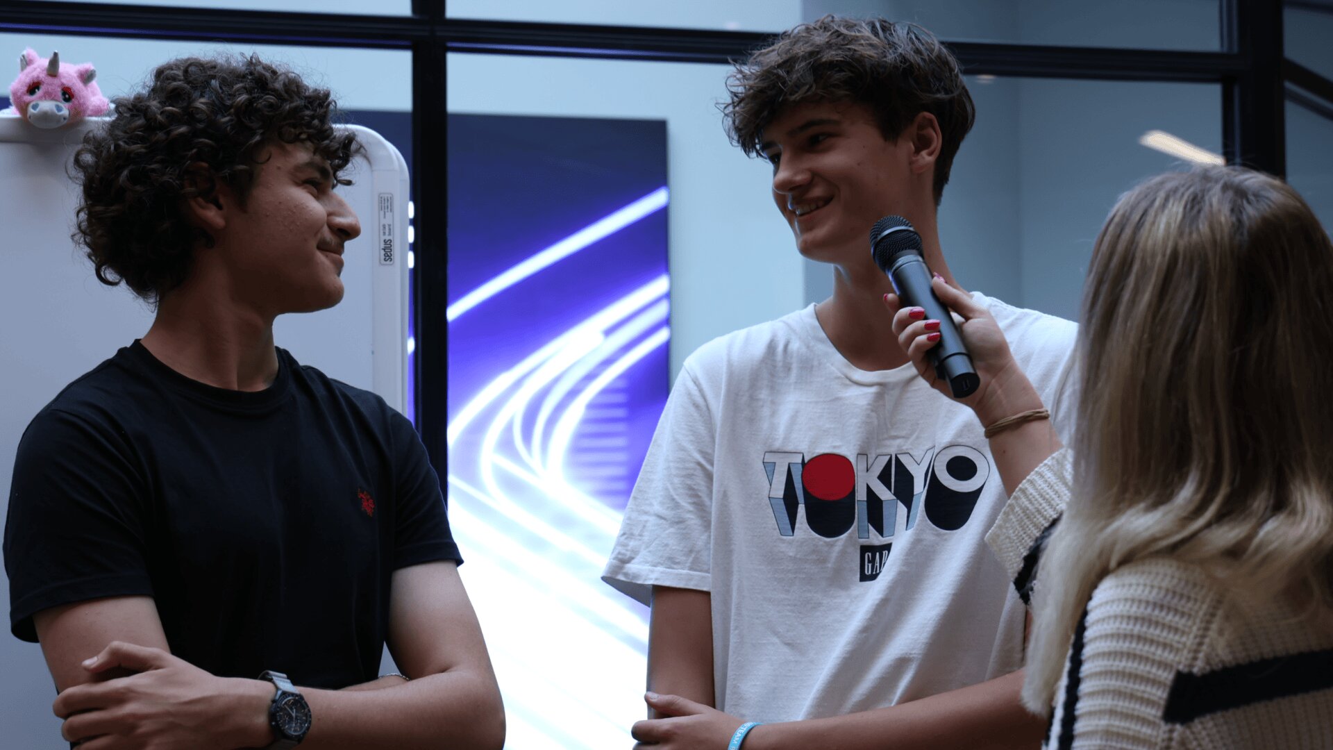 Eine Person hält ein Mikrofon in die Hände von zwei lächelnden Teenagern, die sich gegenüberstehen. Der eine trägt ein schwarzes Hemd, der andere ein weißes Tokio-T-Shirt. Sie stehen in einem Innenraum in der Nähe einer Wand mit einem neonblauen Lichtmuster und einem rosa Plüschtier auf einem Schrank.