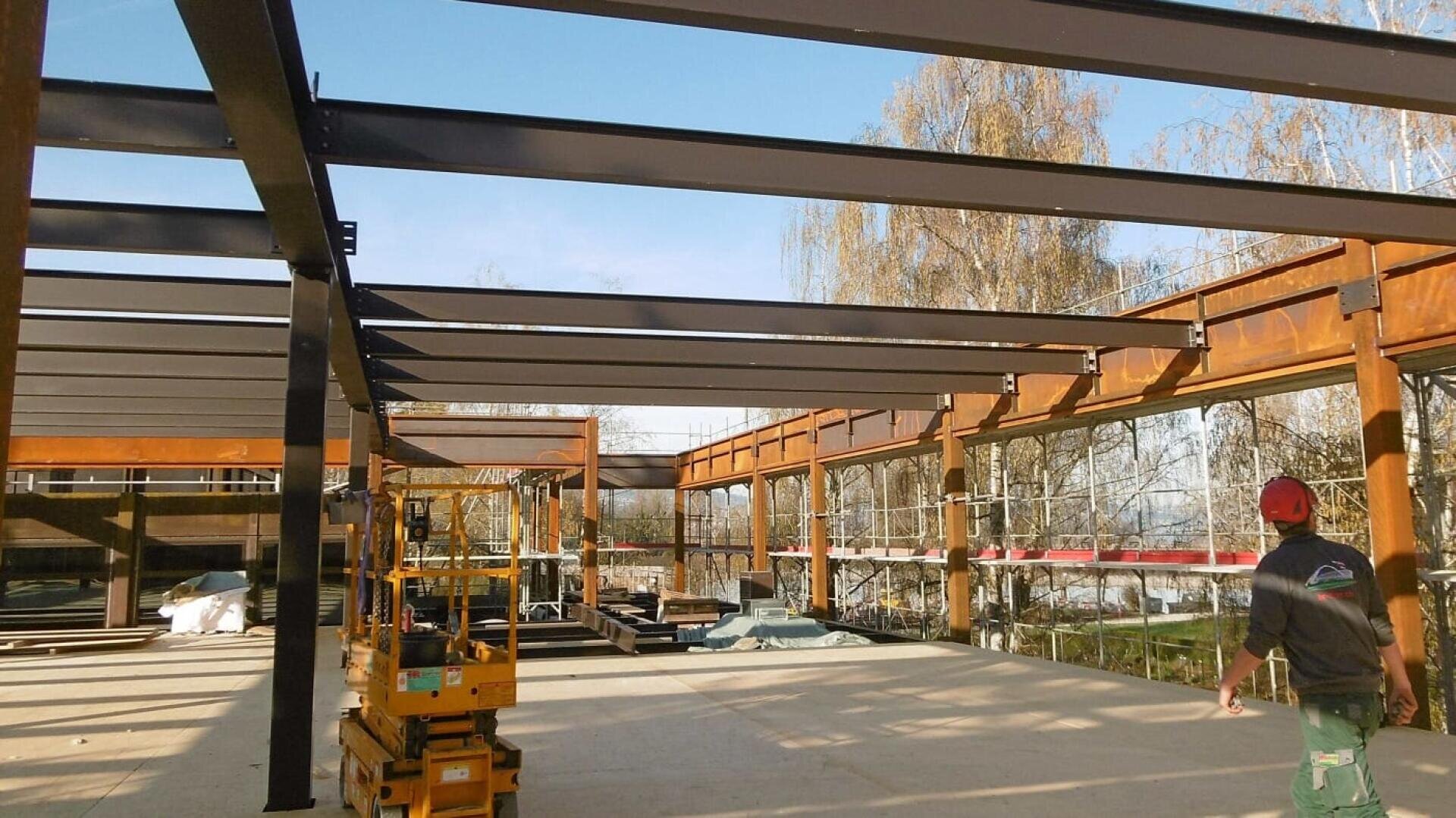 Eine Baustelle mit Stahlträgern, die das Gerüst eines Gebäudes bilden, gelben Gerüsten und Geräten auf dem Betonboden und einem Arbeiter mit Helm und Jacke, der im Vordergrund geht. Draußen sind Bäume zu sehen.