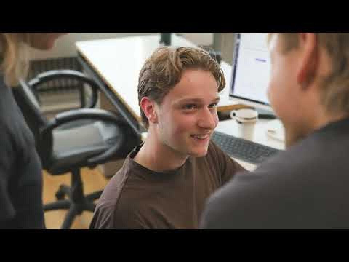 Drei Personen sind in einem Büro um einen Schreibtisch versammelt. Der Fokus liegt auf einem jungen Mann mit hellbraunem Haar, der zwei andere Personen anlächelt, während im Hintergrund ein Computer, eine Tastatur und eine Kaffeetasse zu sehen sind.