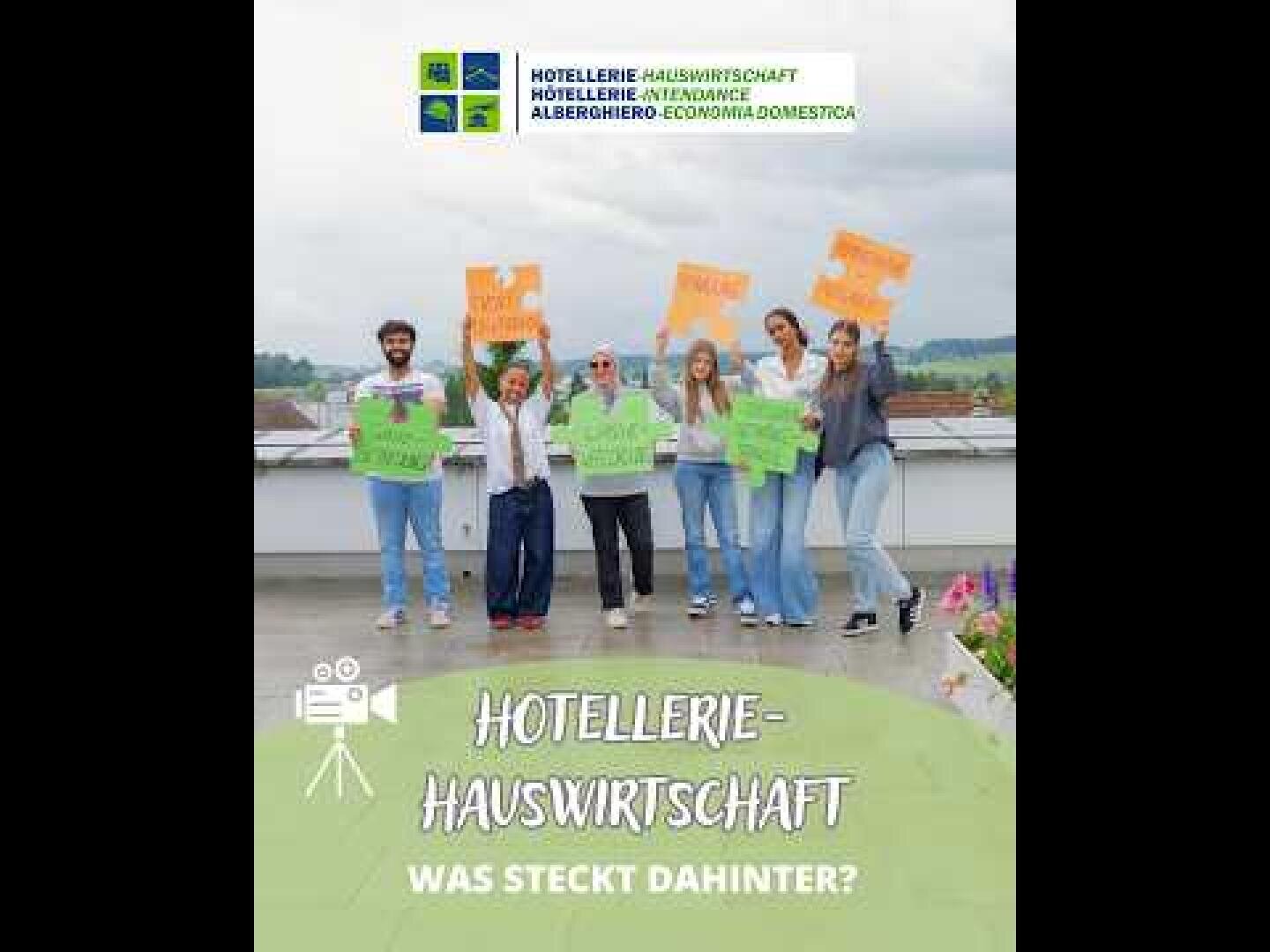 Sieben Personen stehen im Freien und halten bunte Schilder in den Händen. Darauf steht „HOTELLERIE-HAUSWIRTSCHAFT“ und „WAS STECKT DAHINTER?“. Im Hintergrund sind ein klarer Himmel und Grünflächen zu sehen. Das Bild scheint für Berufe in den Bereichen Hotellerie und Hauswirtschaft zu werben.