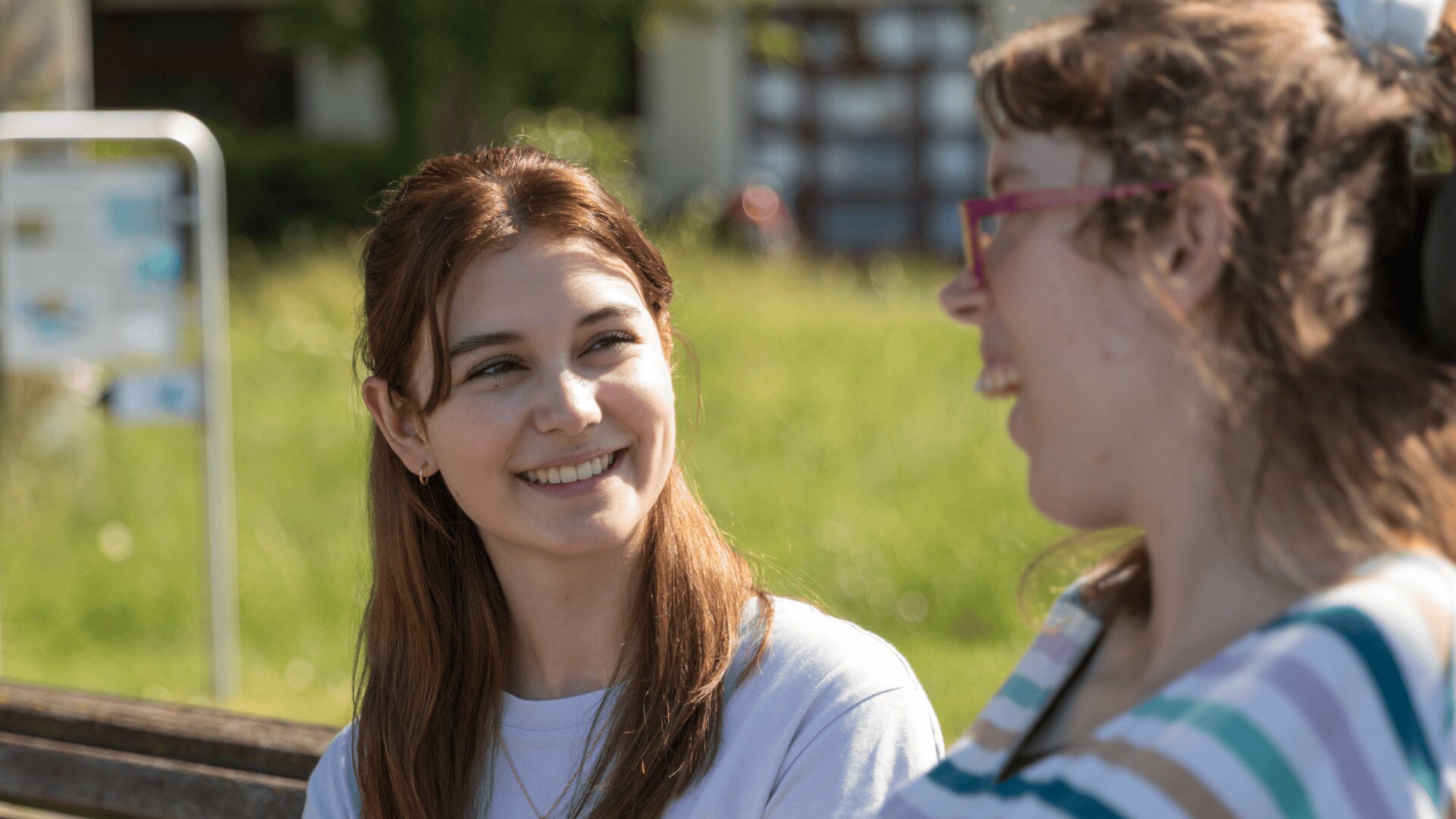 Zwei junge Frauen sitzen an einem sonnigen Tag im Freien auf einer Bank, lächeln und reden miteinander. Die eine Frau hat braunes Haar und trägt ein weißes Hemd, die andere hat eine Brille und trägt ein gestreiftes Oberteil.