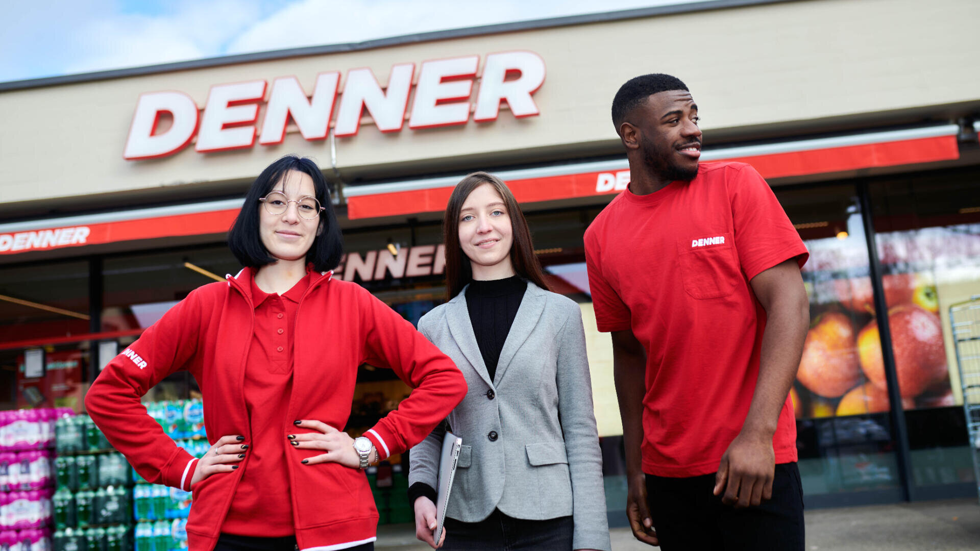 Drei Mitarbeiter stehen lächelnd vor einer Denner-Filiale; zwei tragen rote Denner-Uniformen und einer einen grauen Blazer. Über ihnen ist das Logo des Ladens zu sehen.