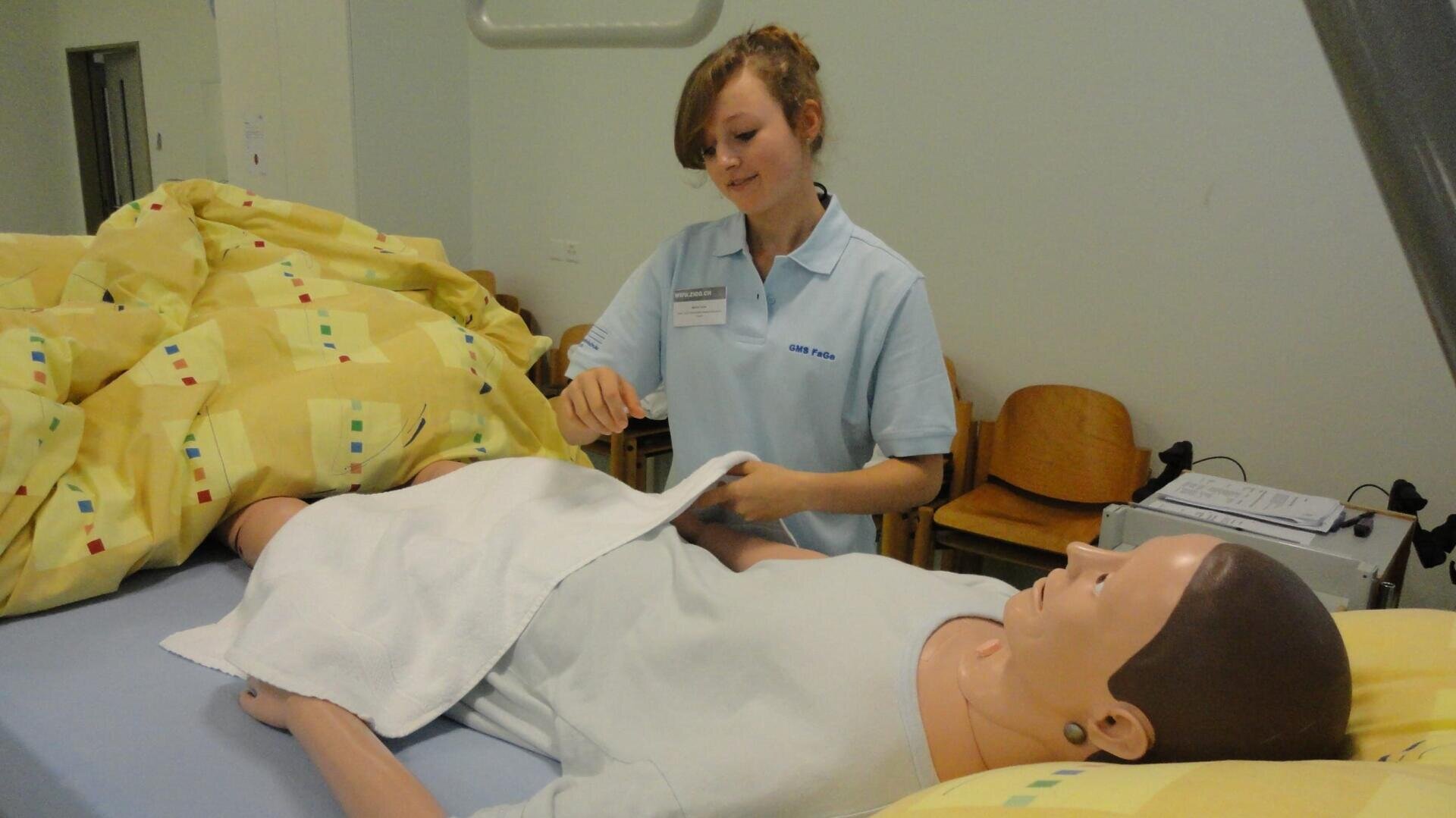 Eine Frau in hellblauer Uniform übt sich in der Patientenpflege, indem sie ein Laken über eine medizinische Übungspuppe legt, die in einem Krankenhausbett mit einer gelb gemusterten Decke liegt.