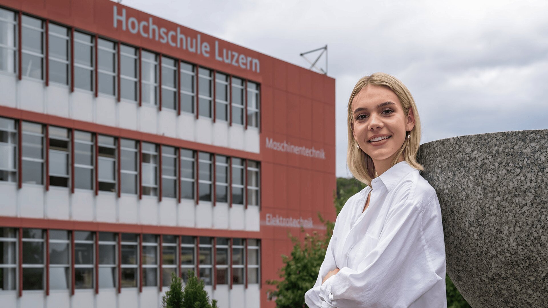 Eine junge Frau mit blondem Haar und weißem Hemd lächelt, während sie draußen vor einem großen Gebäude mit der Aufschrift Hochschule Luzern steht. Der Himmel ist bewölkt und das Gebäude hat viele Fenster.